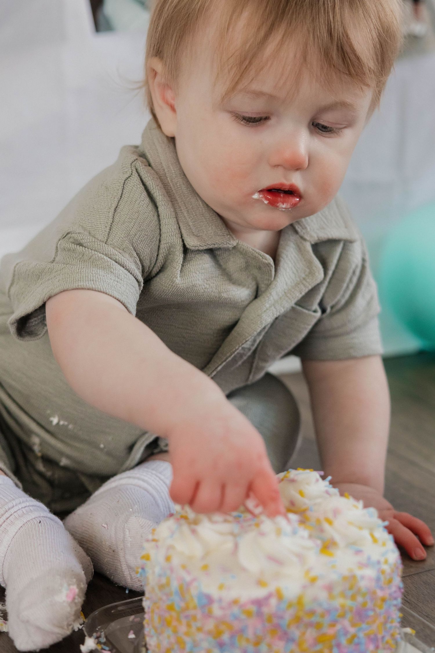 Toddler in sage green outfit reaches into white sprinkle cake during first birthday smash.