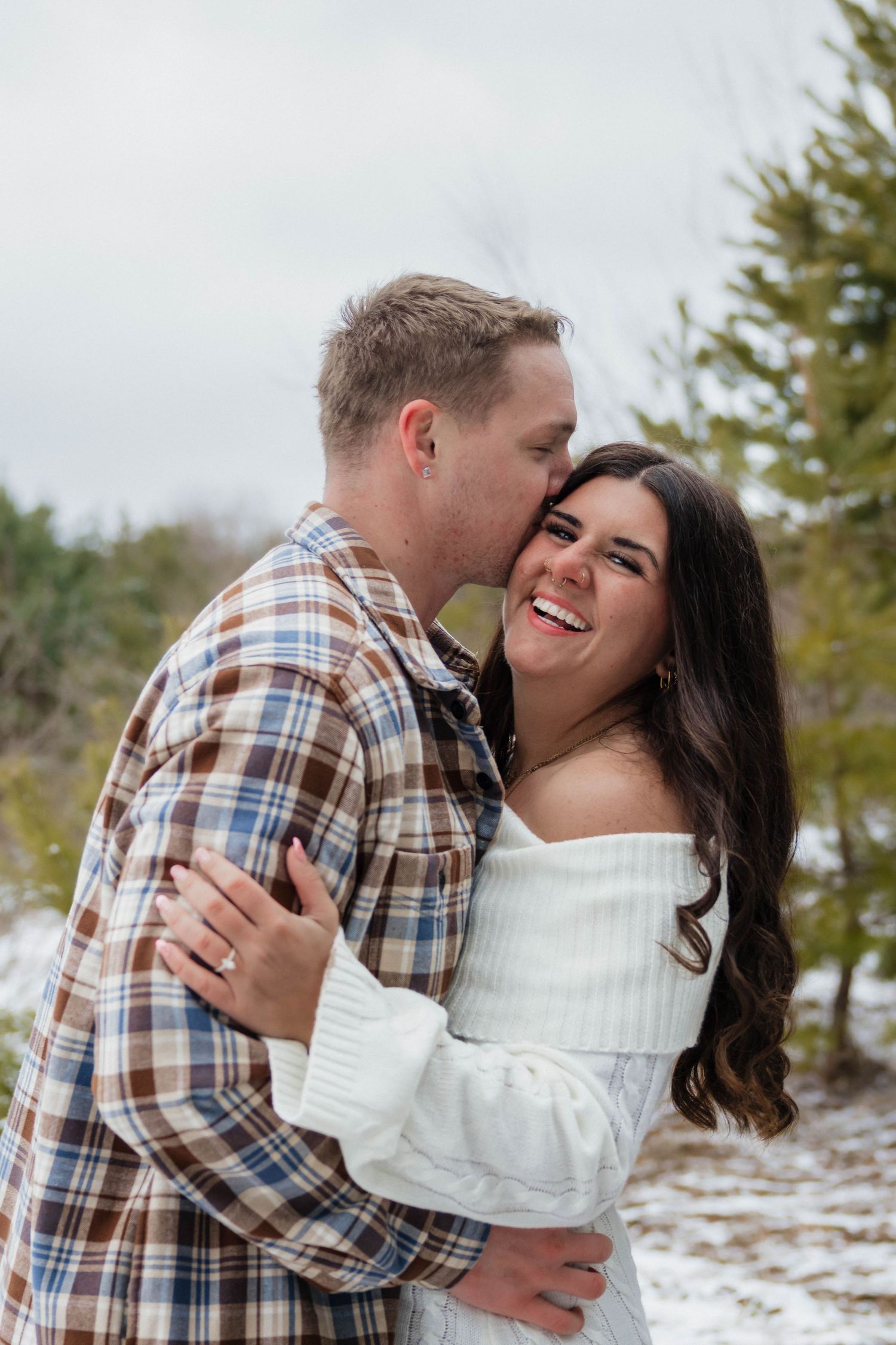 Happy couple sharing a kiss in snowy evergreen forest, woman laughing in white off-shoulder sweater.