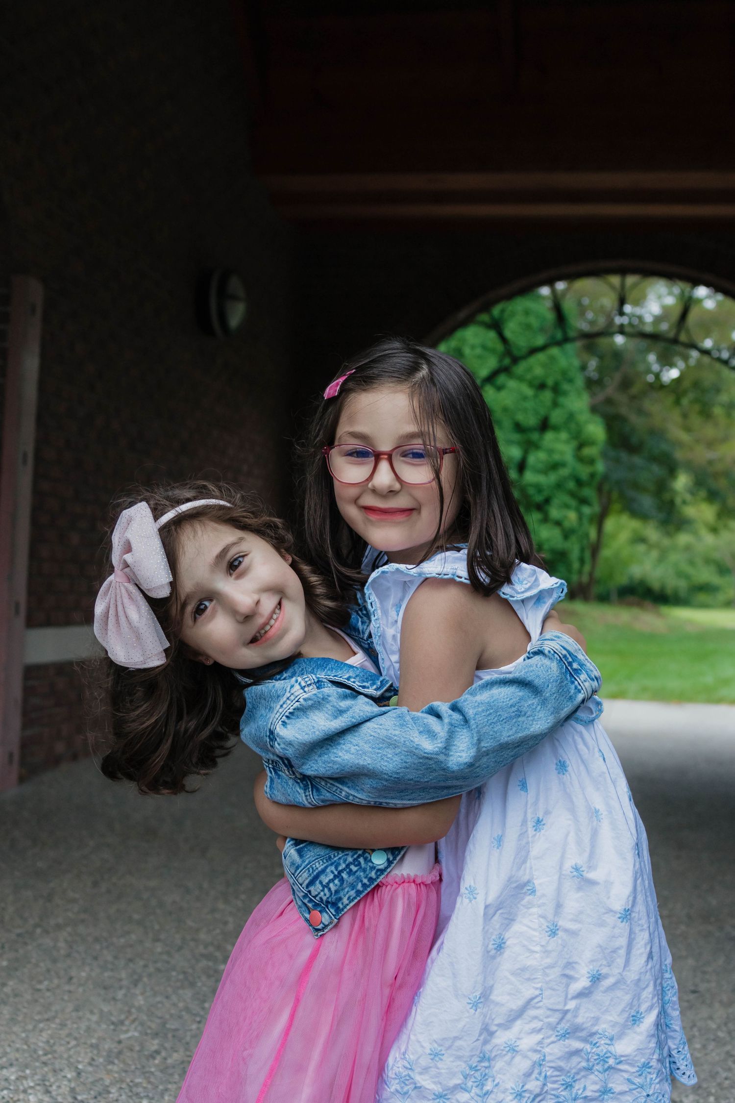 Two young girls hugging and smiling under an archway, one in pink tutu and one in a blue dress with glasses.