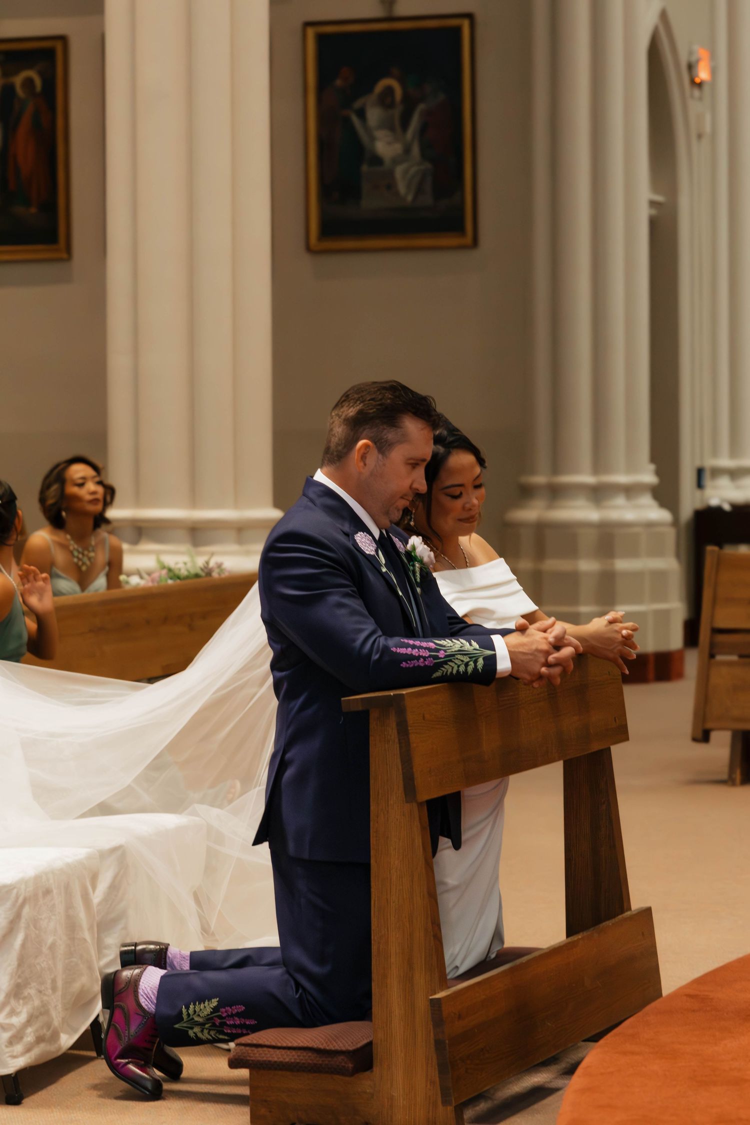 Bride and groom kneel together in prayer at wooden pew during their church wedding ceremony.