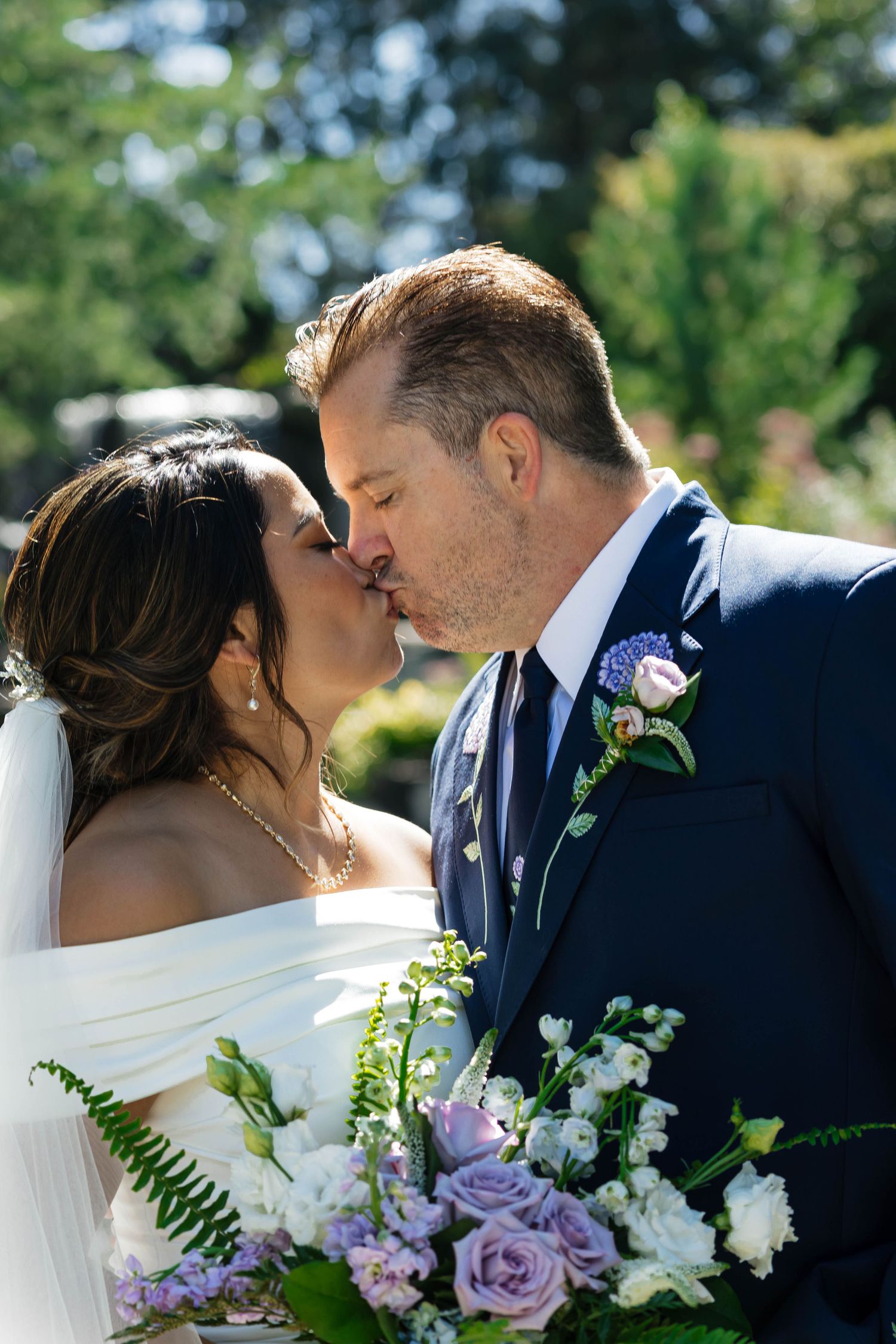 Bride and groom share a romantic first kiss at outdoor wedding ceremony with purple and white floral bouquet in foreground.