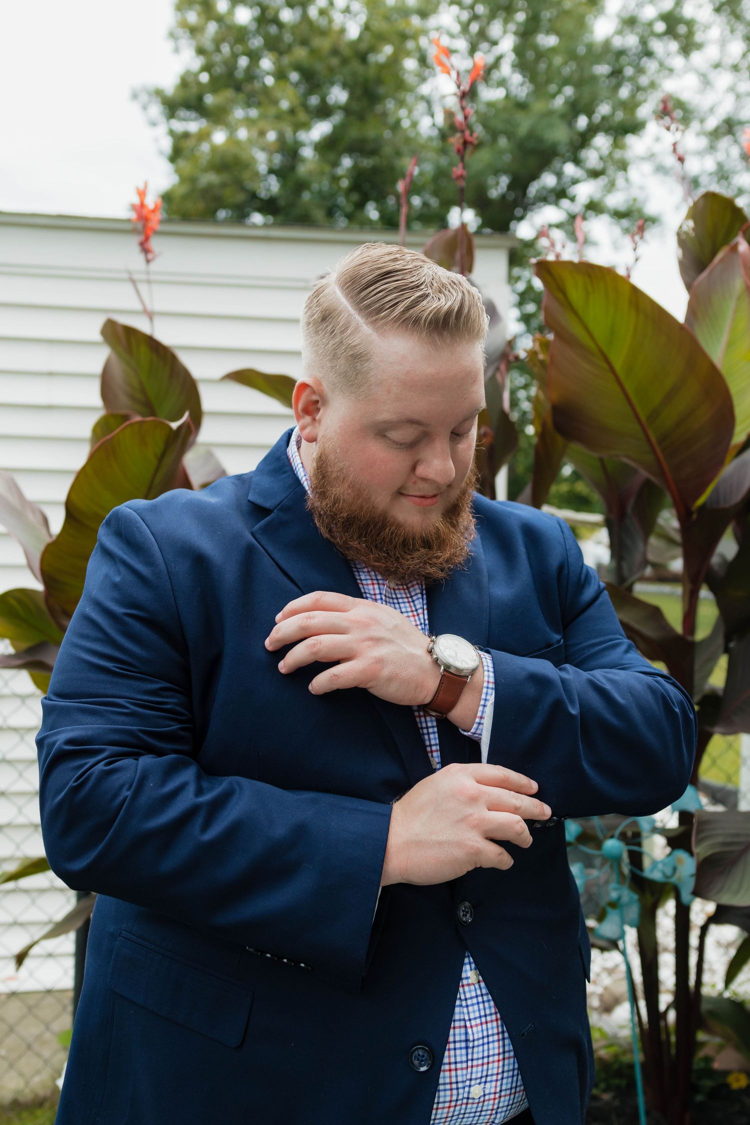 Bearded groom in navy blue suit adjusting watch cufflink in outdoor garden setting.