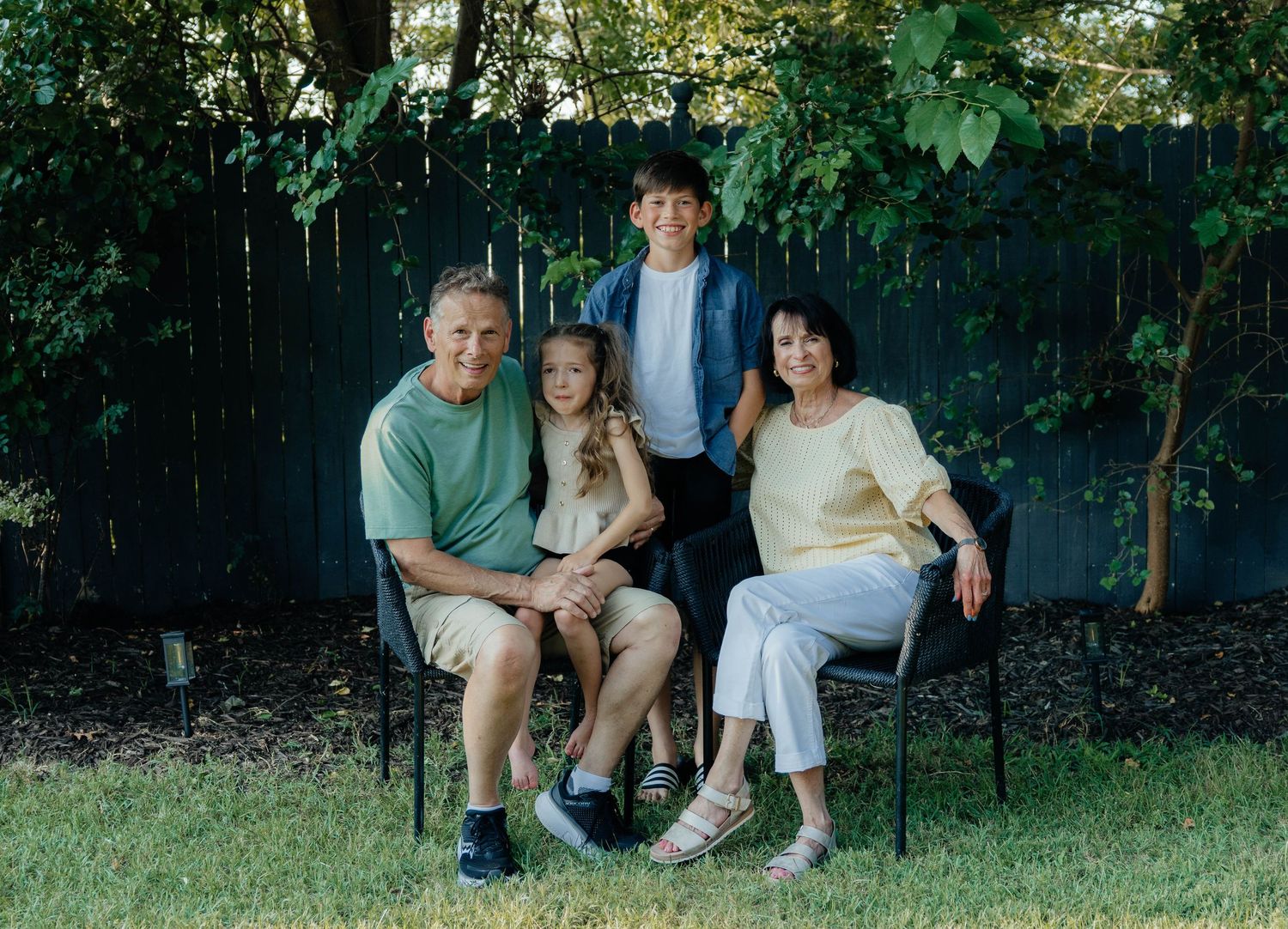 Grandparents sitting in chairs outdoors with two grandchildren posing in a backyard.