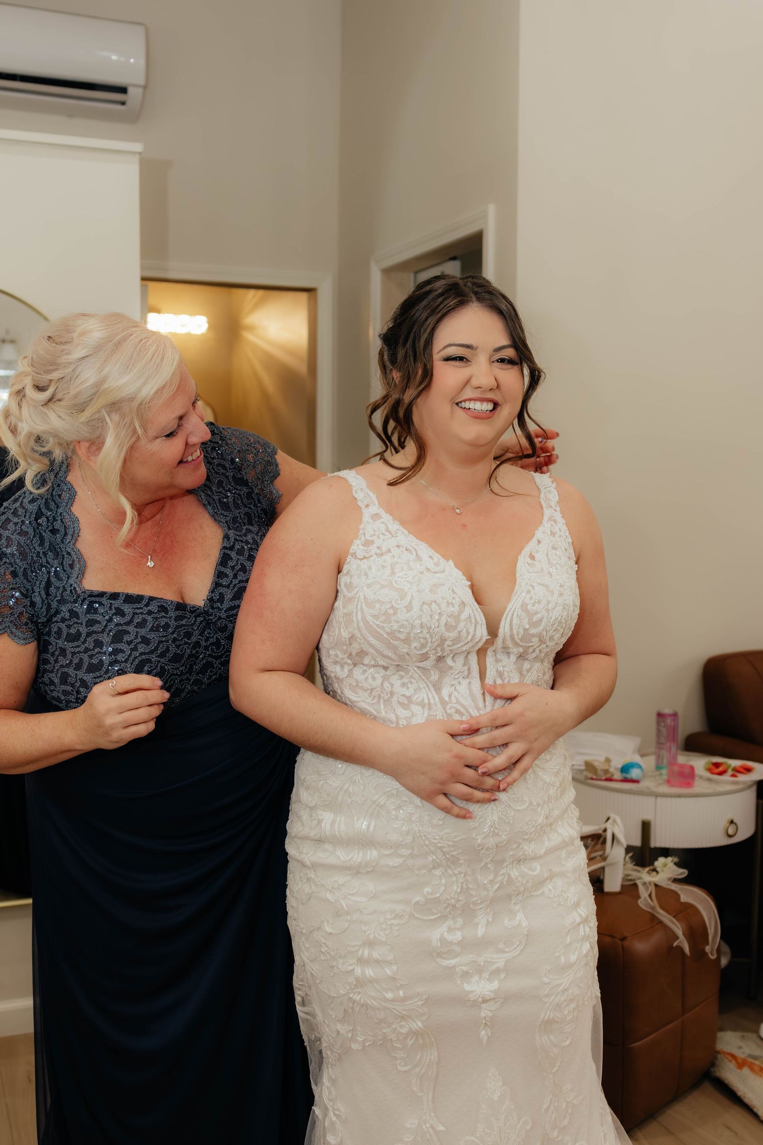 Joyful bride in lace wedding gown being helped by older woman in navy dress indoors.
