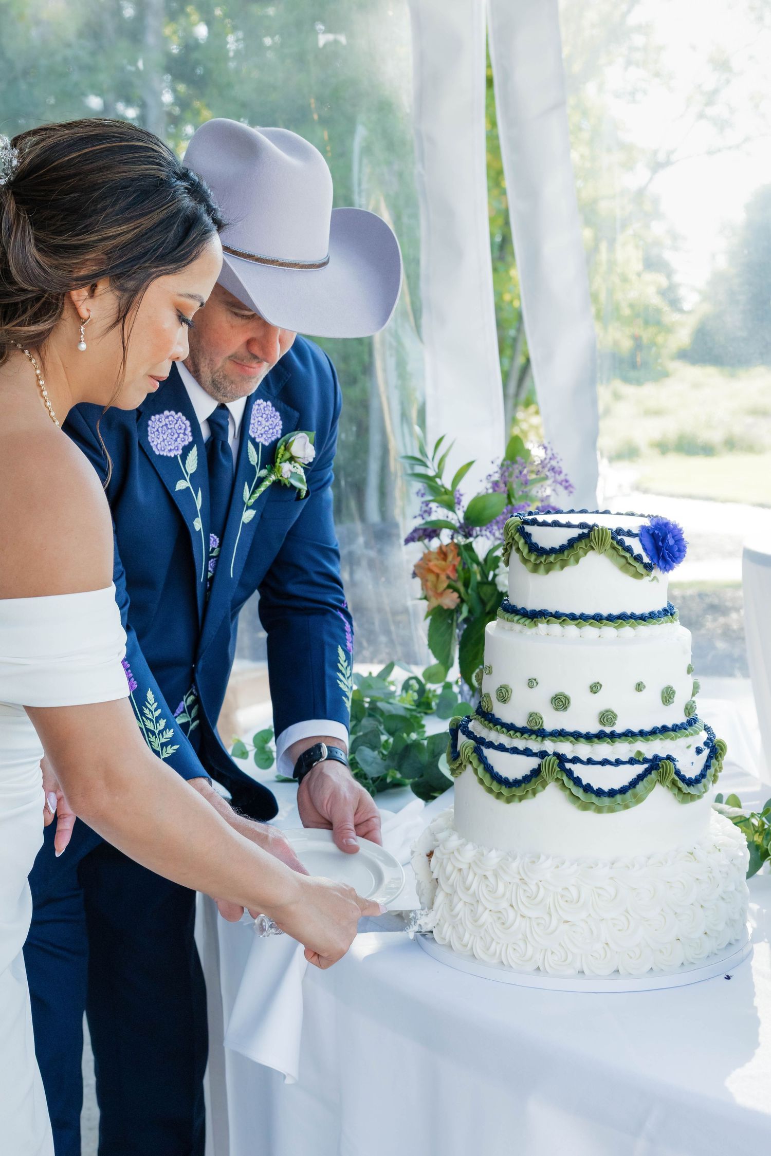 Bride and groom in navy suit and cowboy hat cut three-tier white wedding cake with green polka dots.