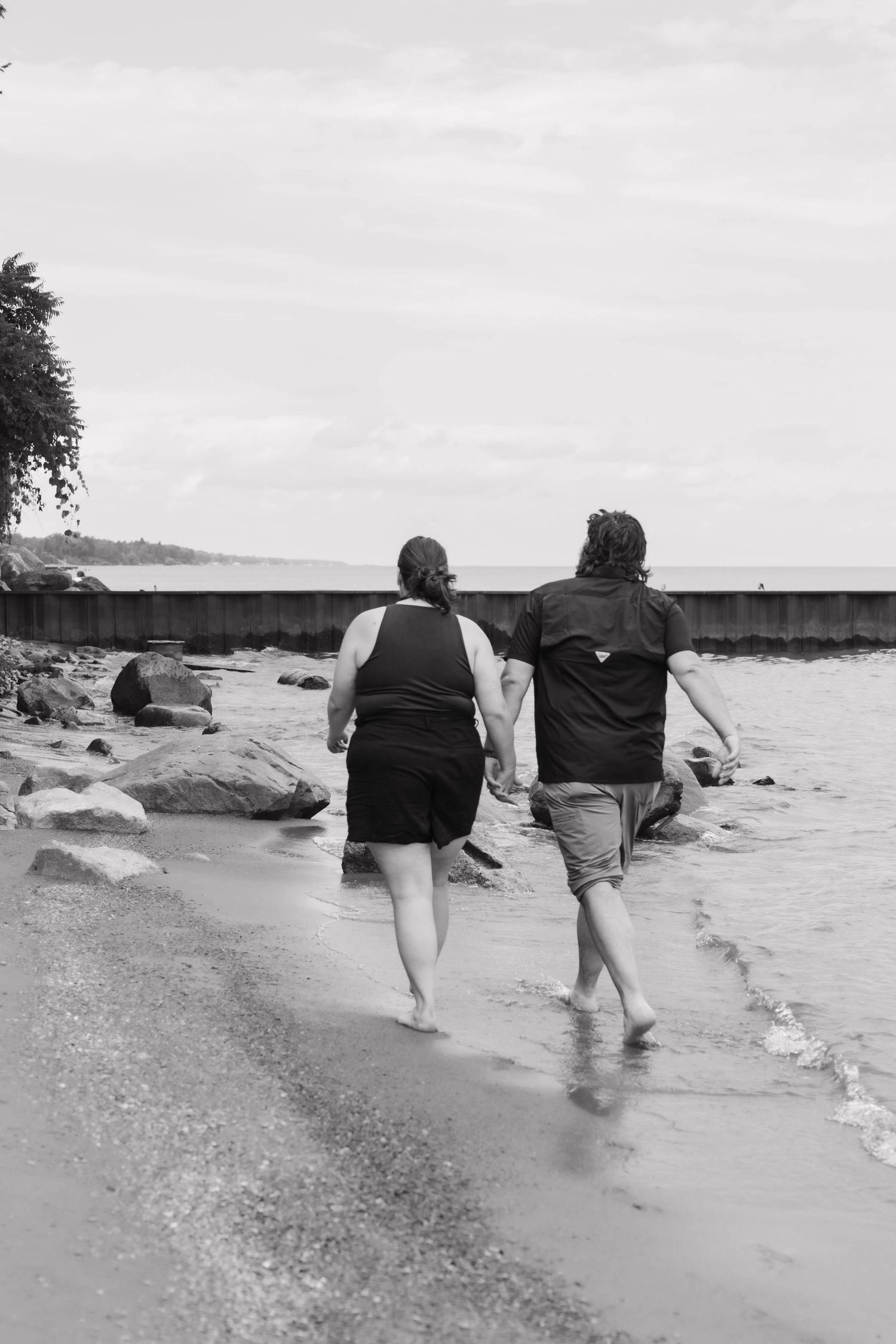 A black and white photo of a couple walking barefoot along a rocky shoreline together.