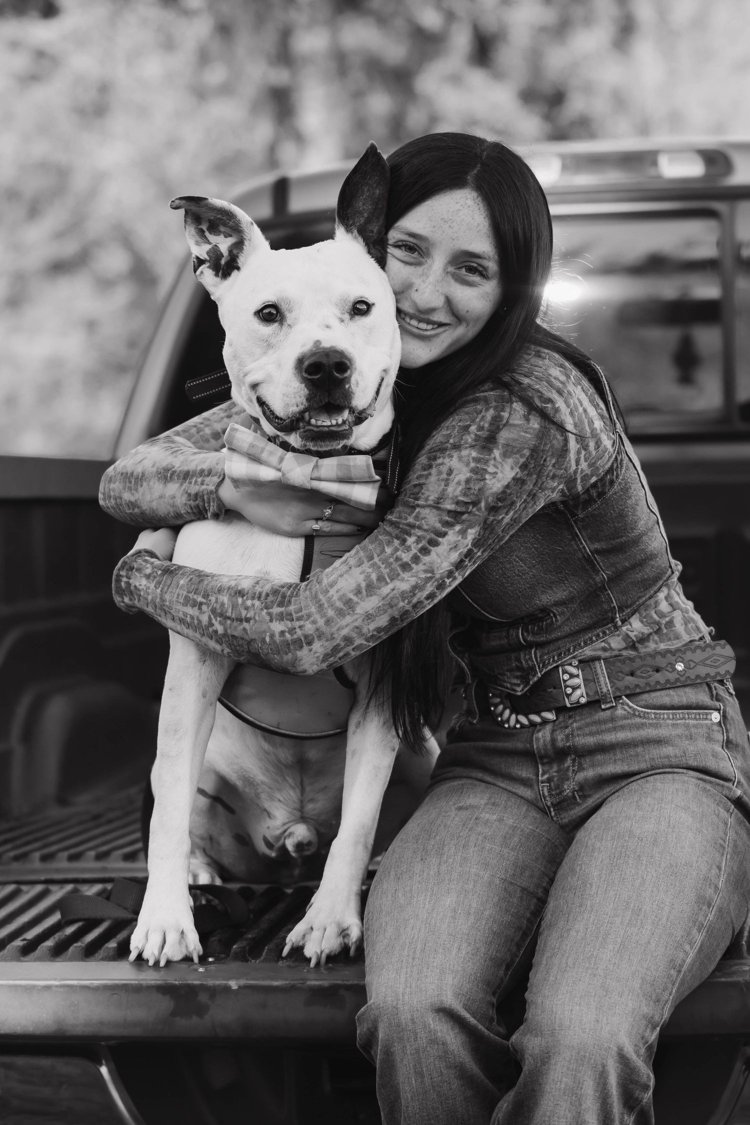 Black and white photo of smiling woman hugging a happy white pitbull dog on a truck tailgate.