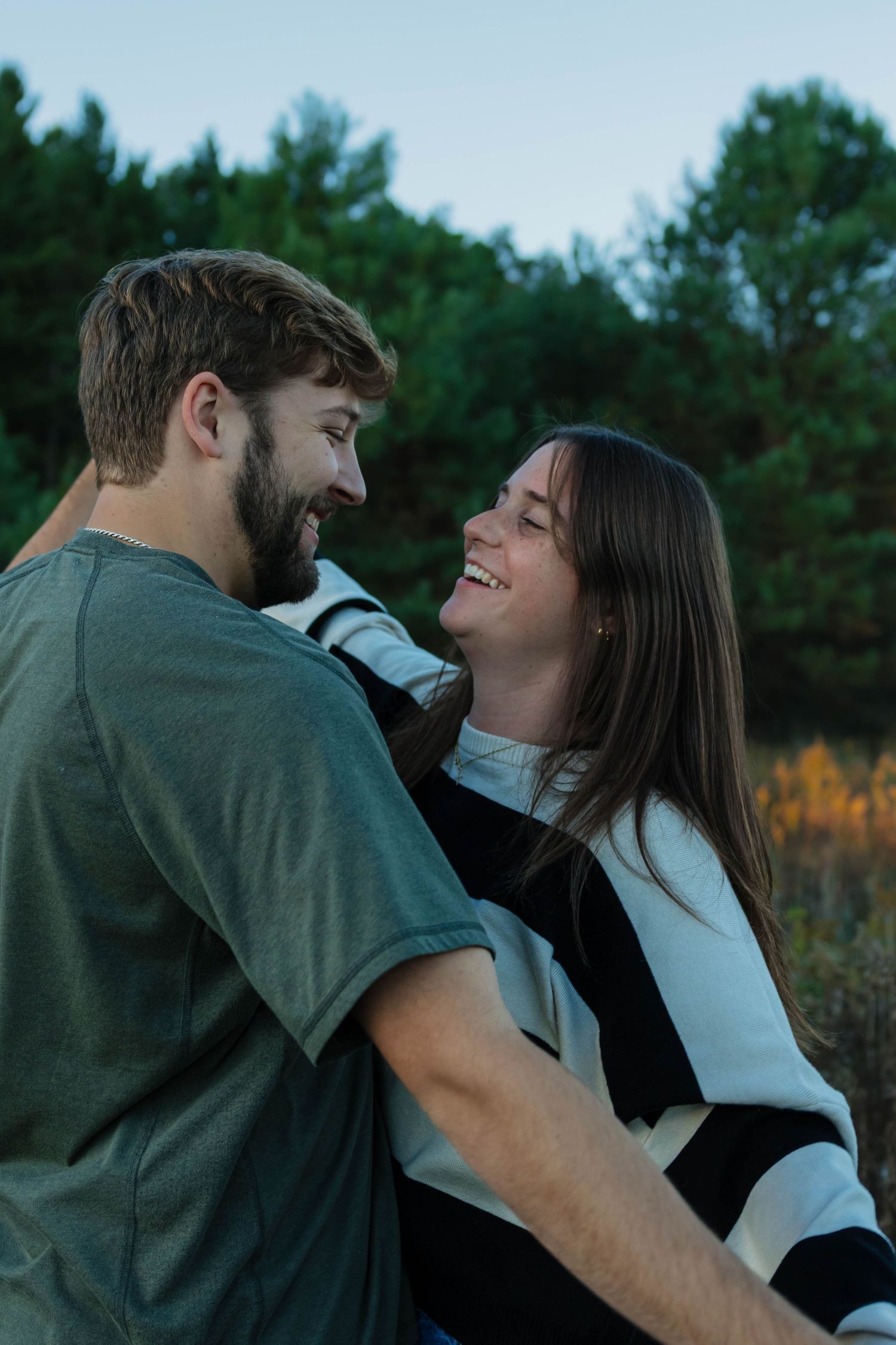 Happy couple laughing and embracing outdoors at dusk surrounded by lush green trees.