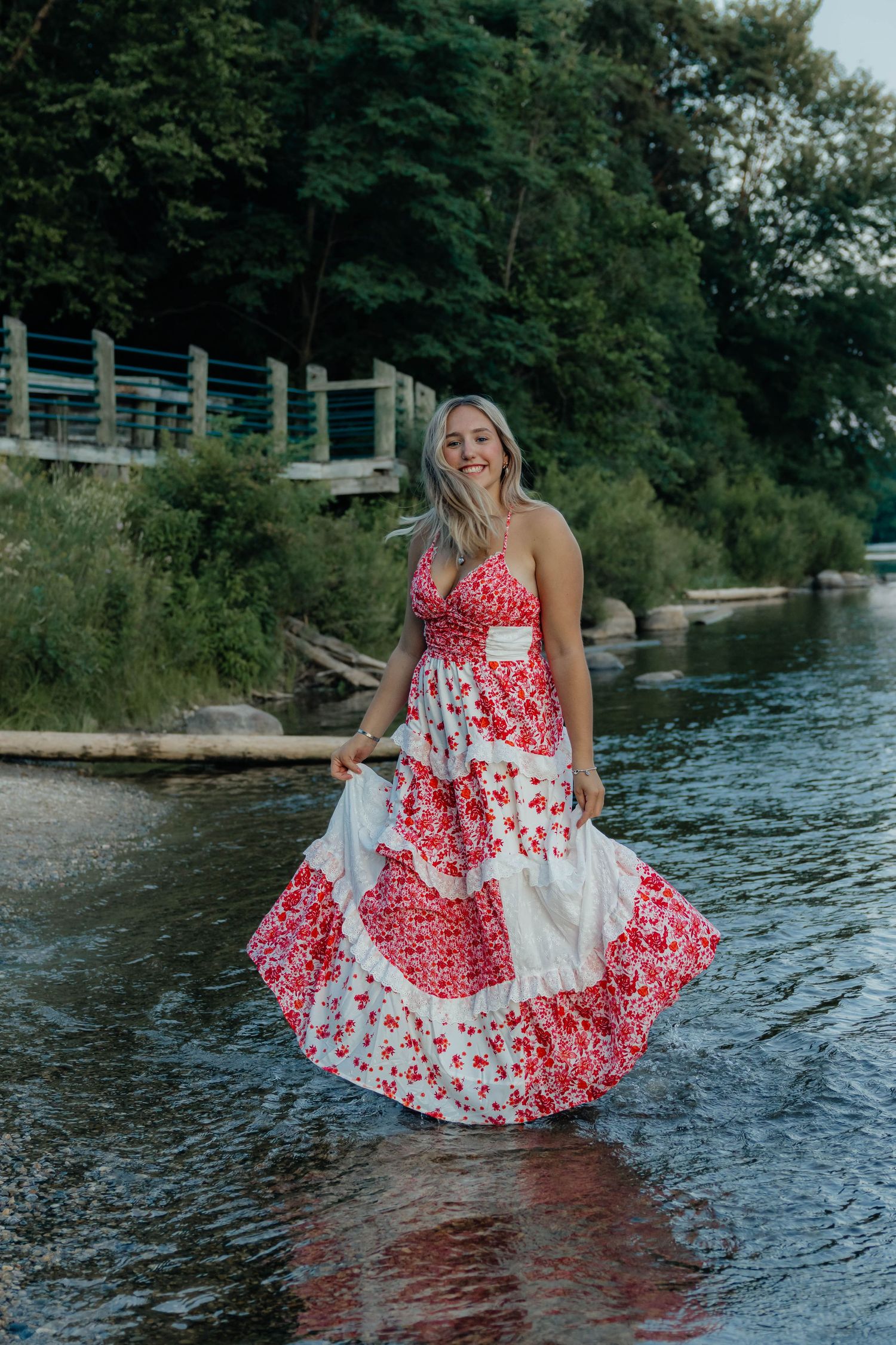 Laughing blonde woman in red floral maxi dress twirling in river water at golden hour.