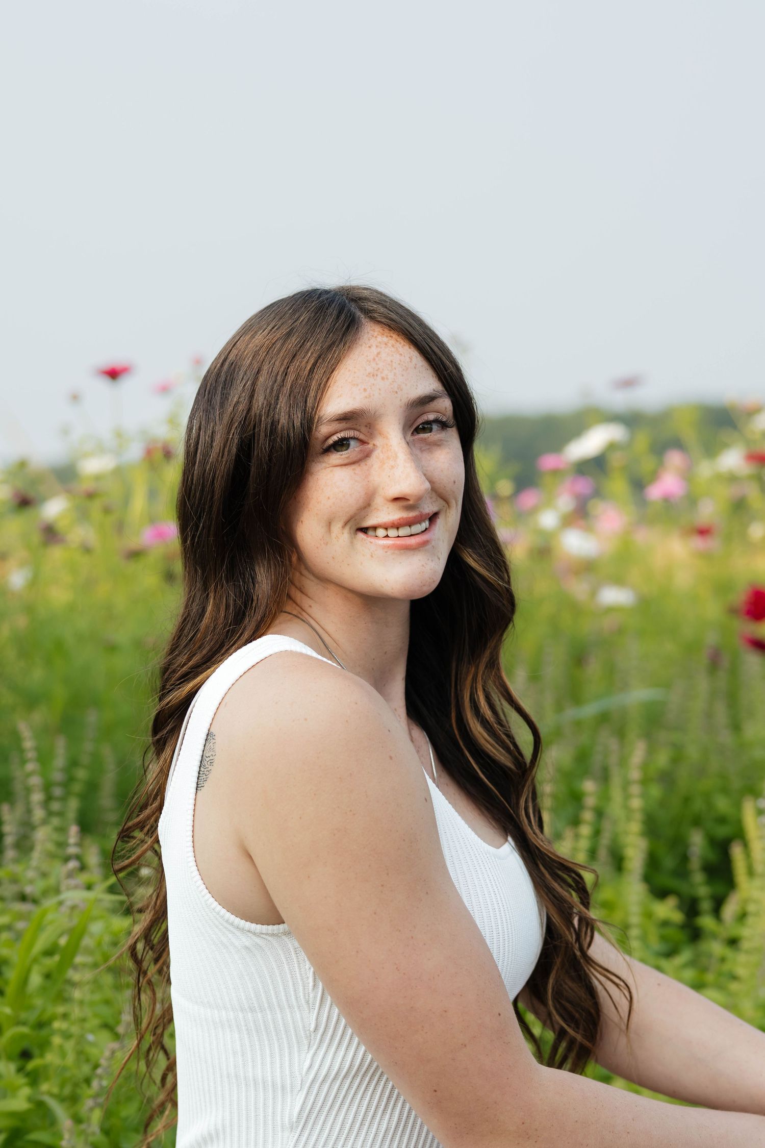 Smiling teenage girl with freckles and long brown hair in white top among colorful wildflowers.