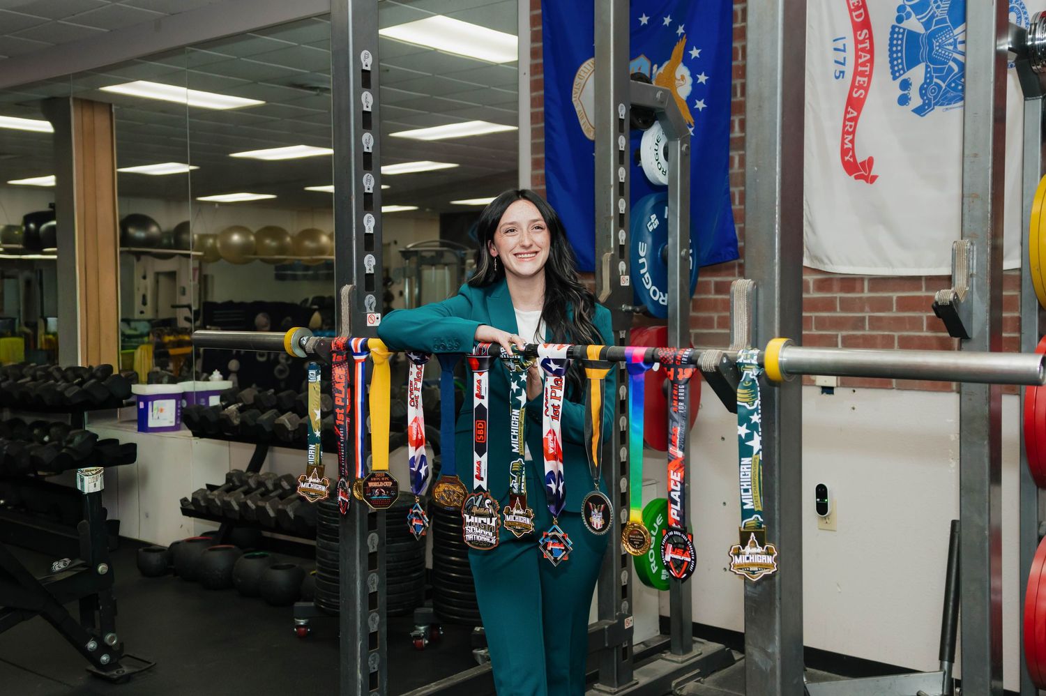 Young woman in teal outfit proudly displaying collection of colorful medals in school weight room.