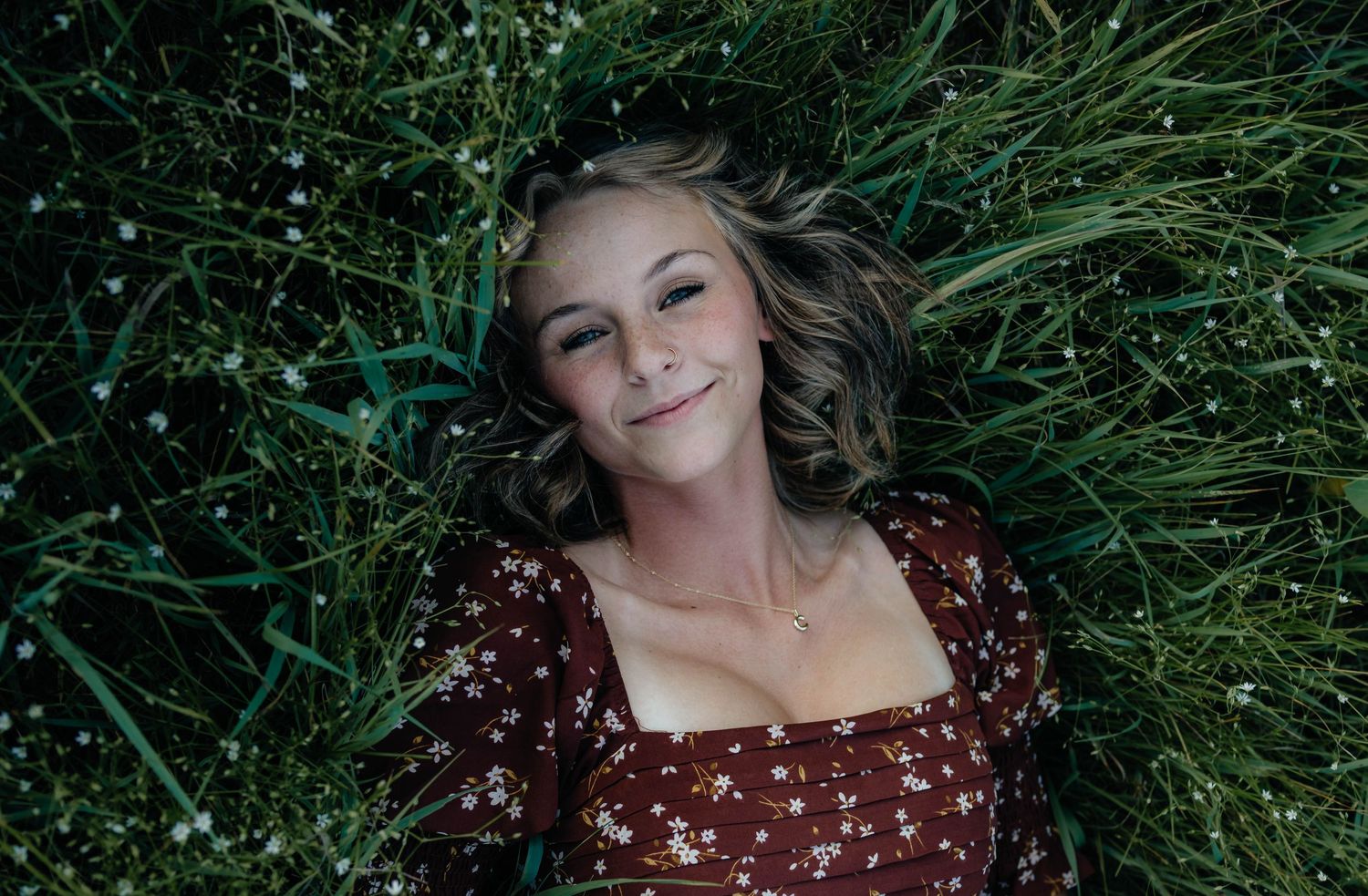 Young woman in a floral dress lying in green grass, smiling up at the camera with a relaxed expression.