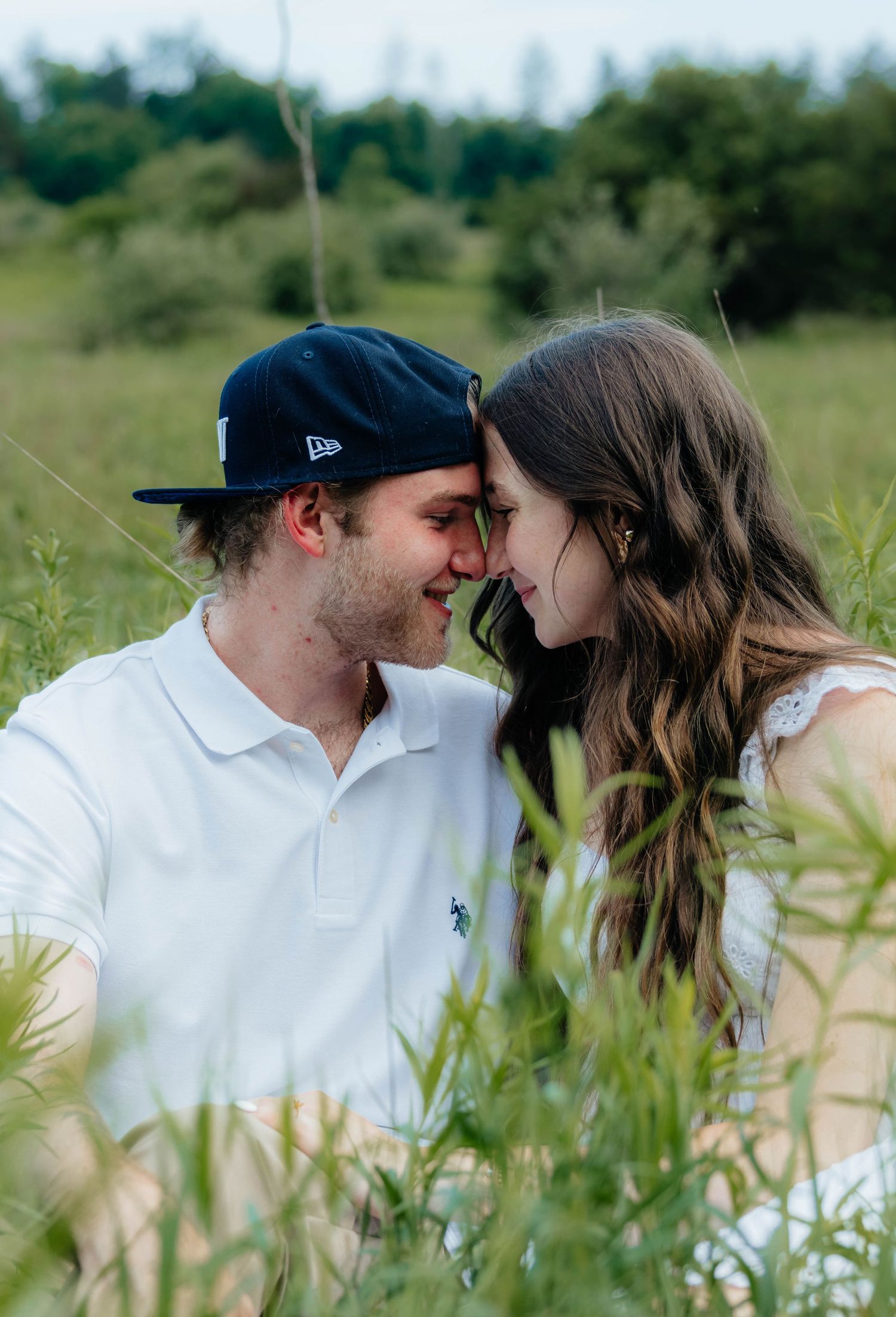 Romantic couple touching foreheads and smiling while sitting together in tall green grass.