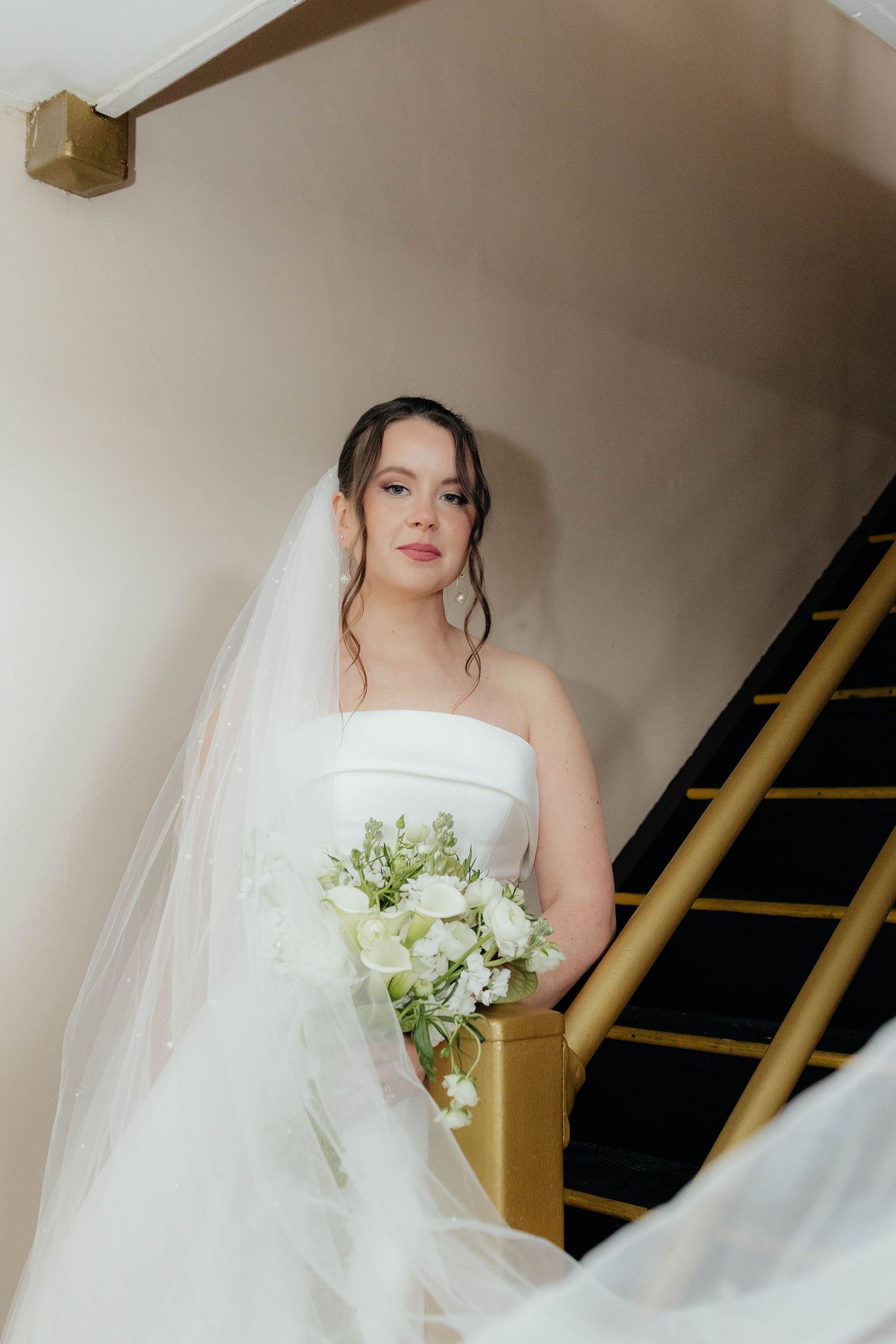 Bride in strapless white gown and veil holds white bouquet on staircase.