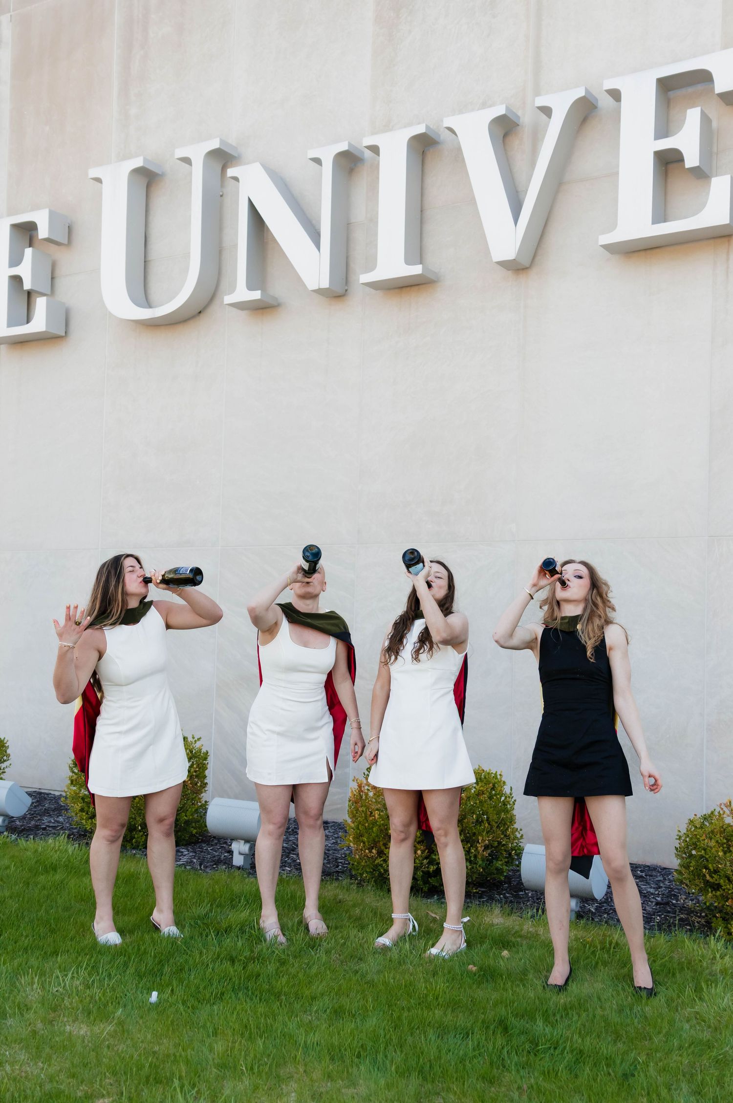 Four young women in white and black dresses playfully drink from bottles in front of a university sign.
