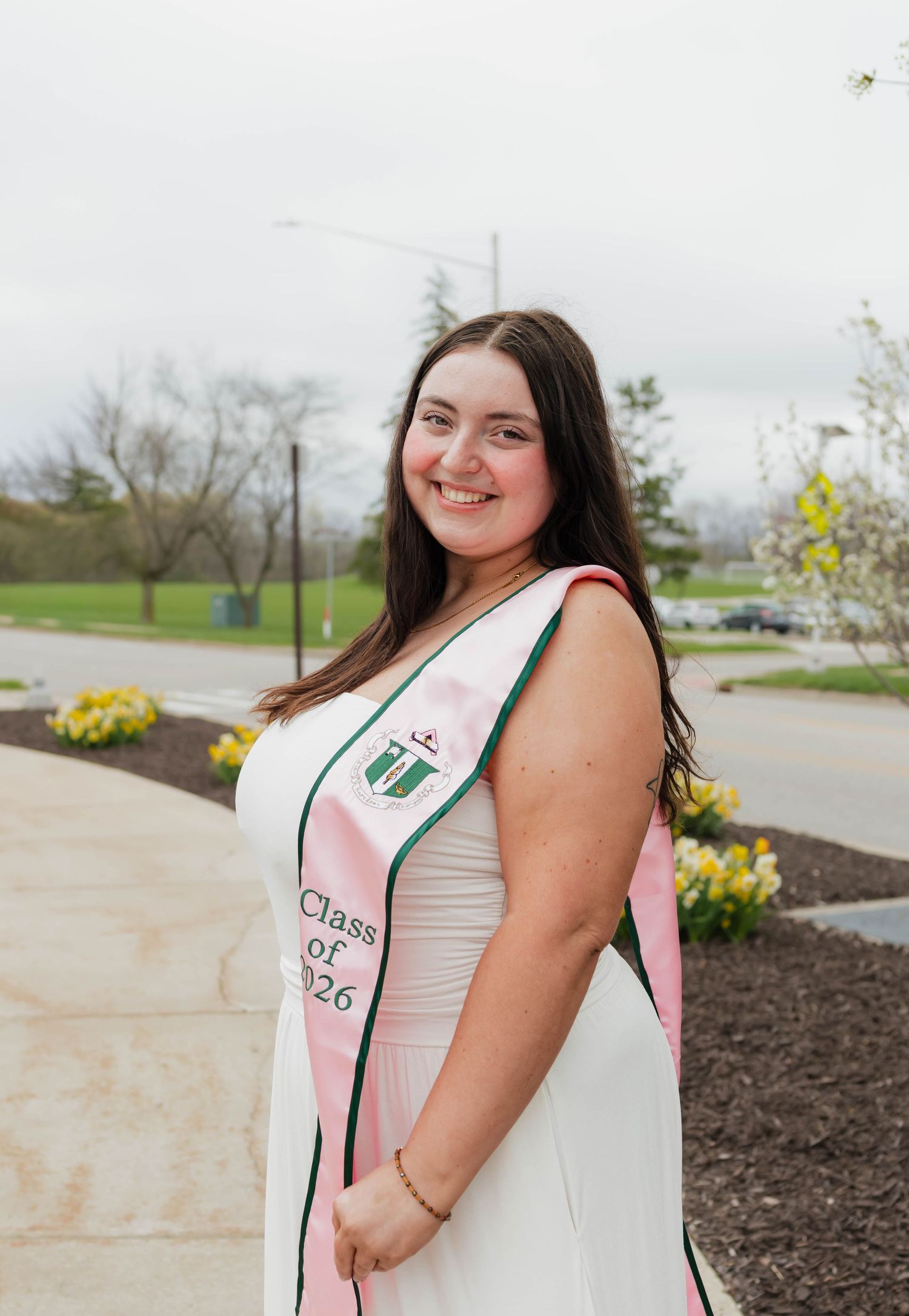 A smiling graduate wearing a pink Class of 2026 sash over a white dress poses outdoors in spring.