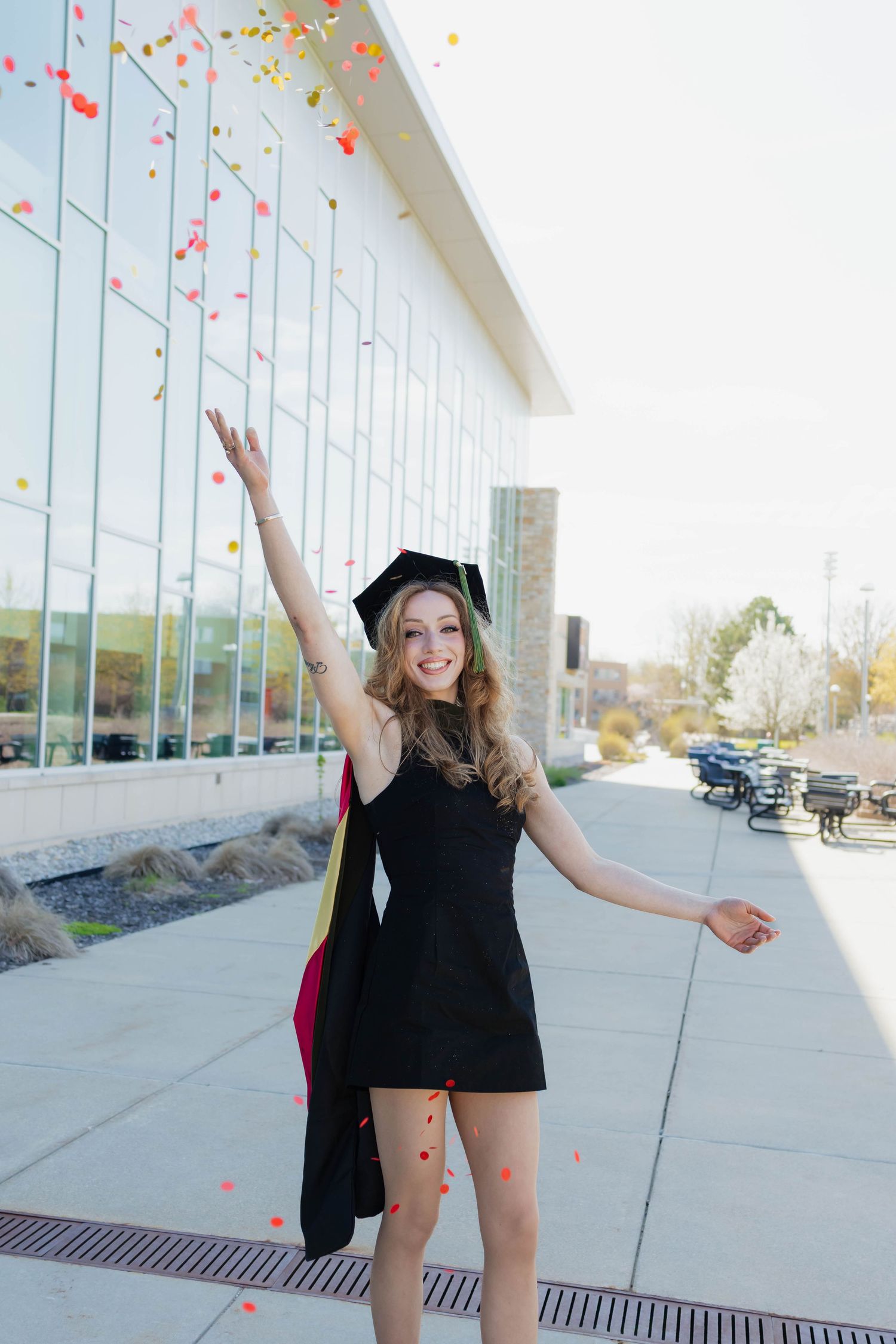 Excited blonde graduate in black dress tosses colorful confetti outside glass building.