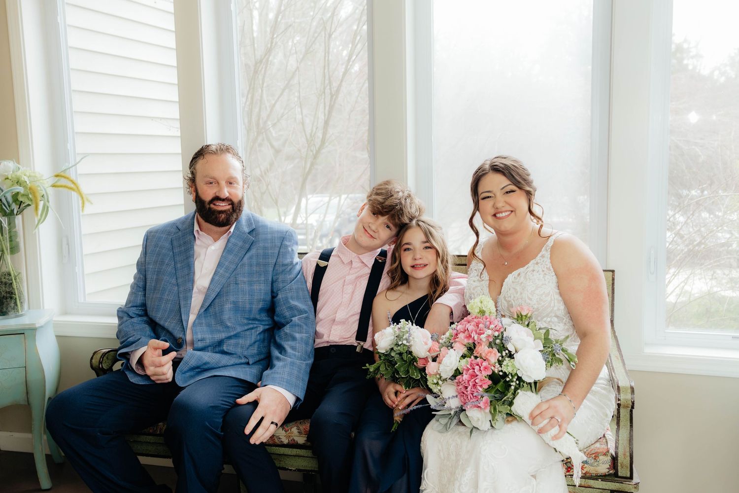 Bride holding pink bouquet poses with groom in blue suit and two children by sunlit window.