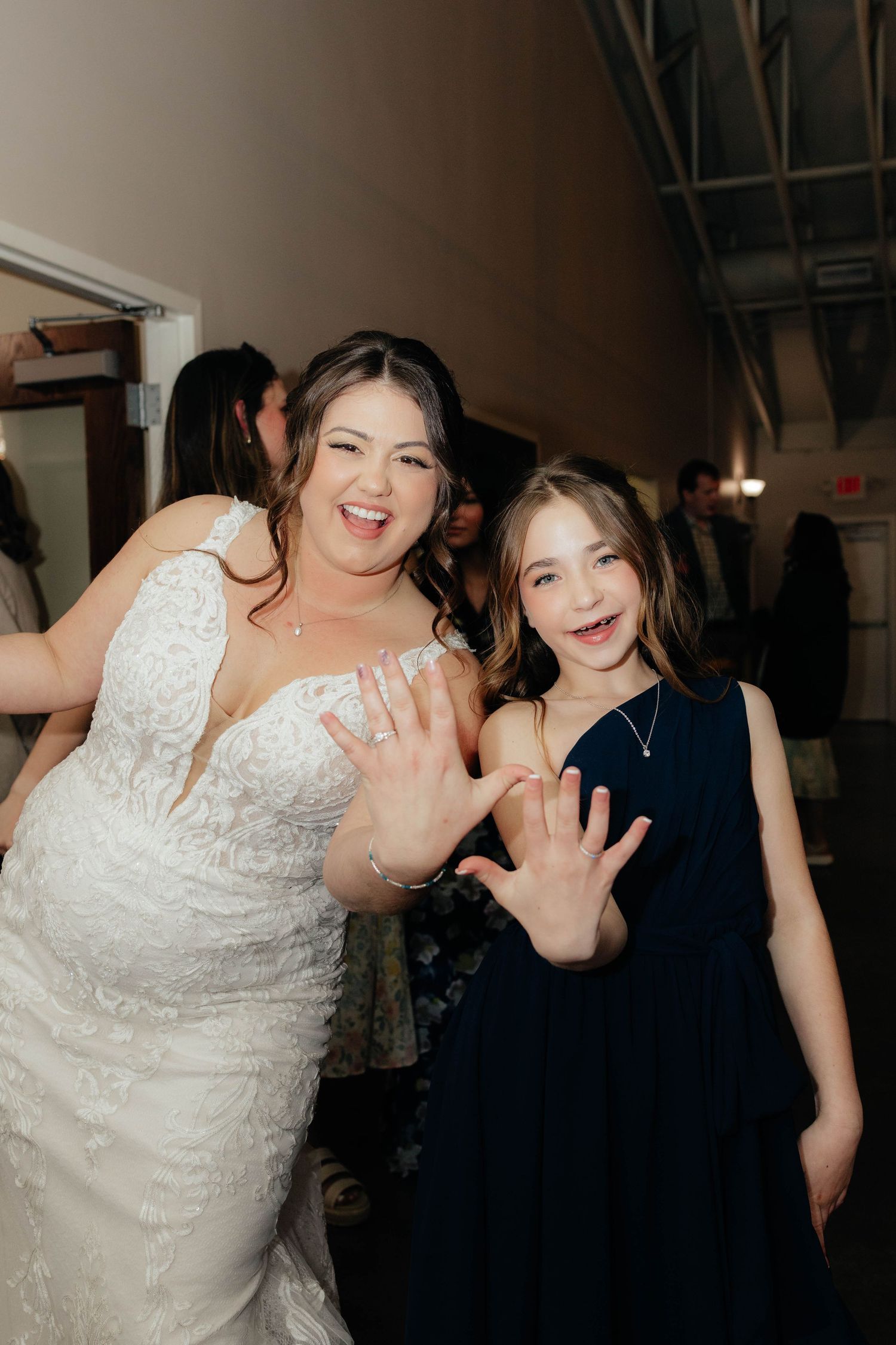 Bride in lace wedding gown and young girl in navy dress joyfully showing off rings at wedding reception.