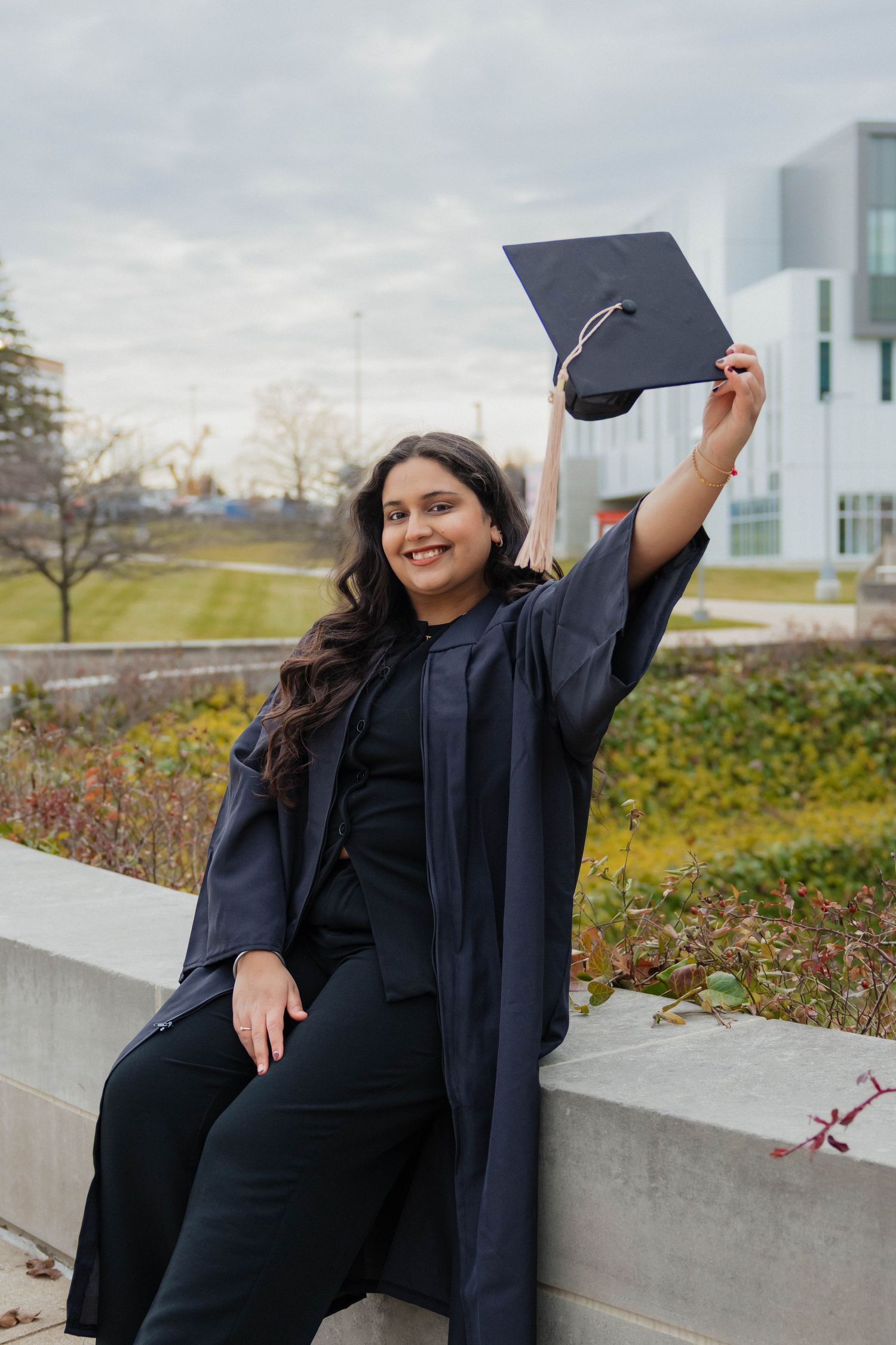 Joyful graduate in navy gown holding mortarboard cap up high while sitting on a campus wall.