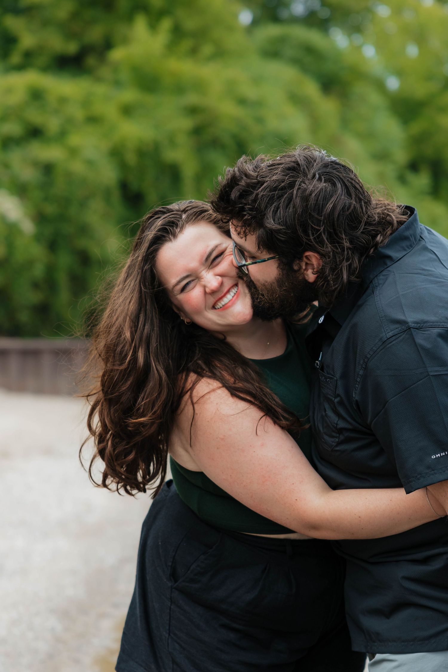 A couple shares a joyful embrace outdoors, with the man kissing the woman's cheek as she laughs.