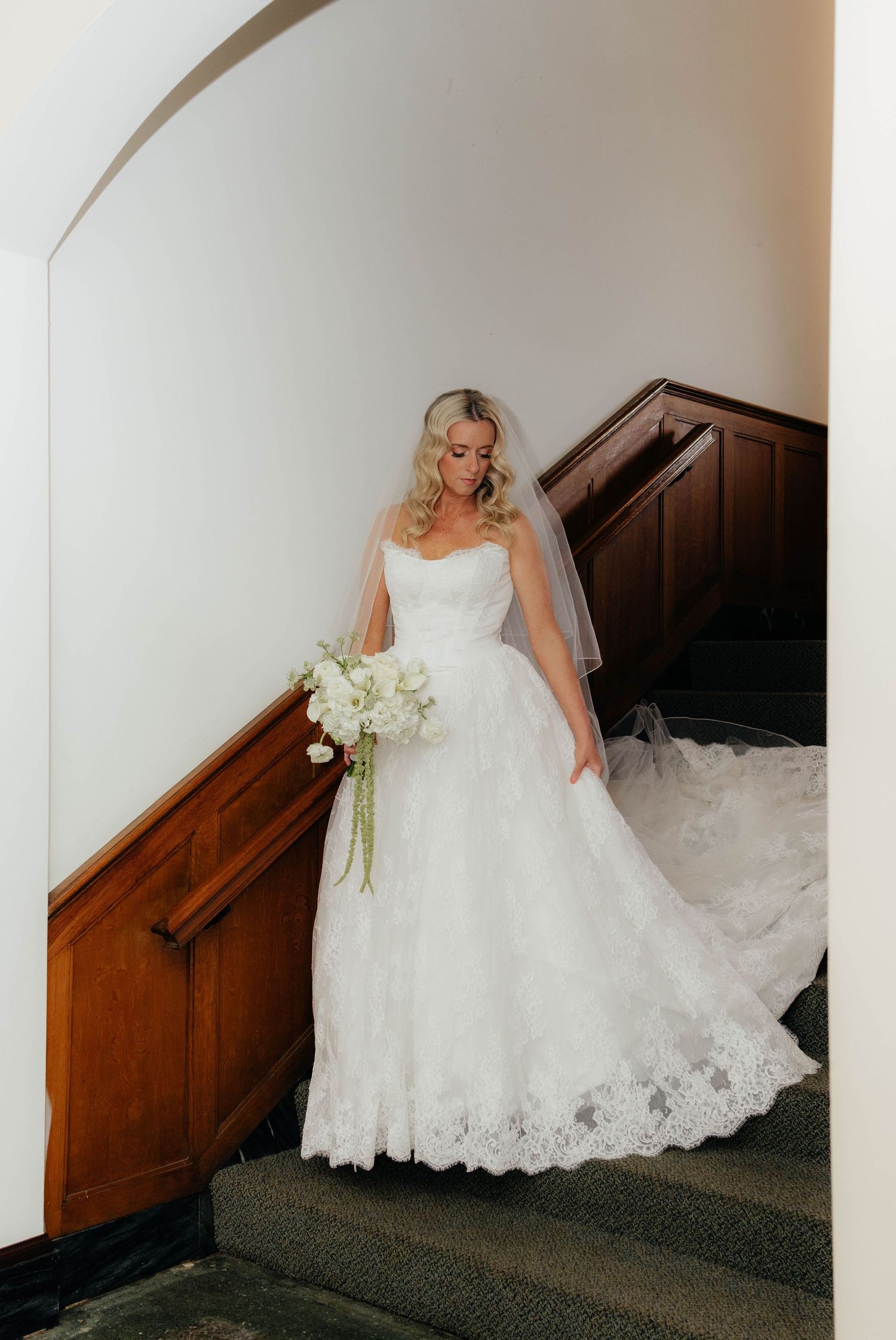 Bride in strapless lace ball gown with veil descending elegant wooden staircase with bouquet.