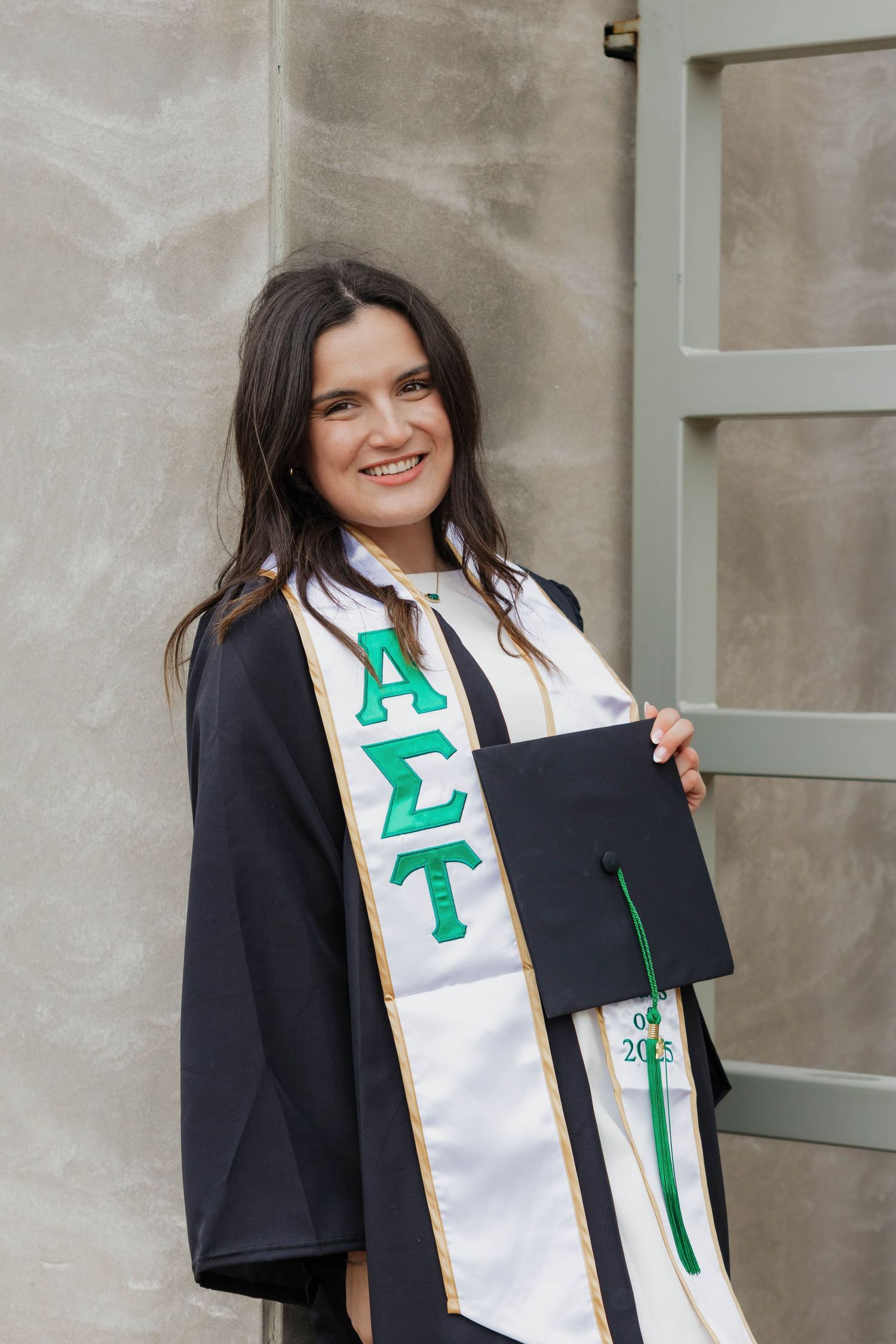 Smiling woman in graduation gown holding cap with Alpha Sigma Tau sorority stole.