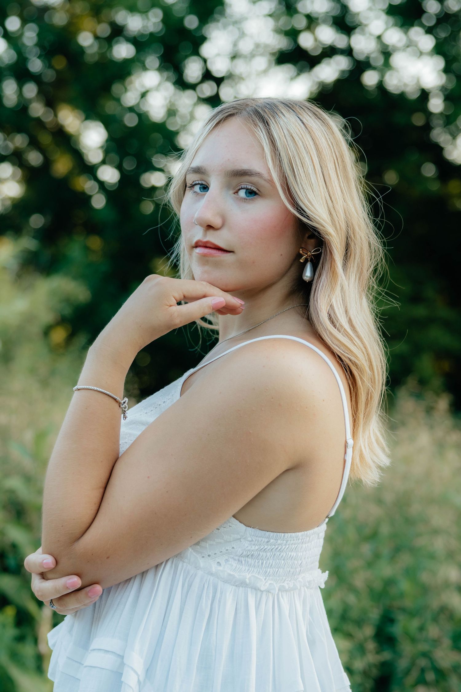 Blonde teen with blue eyes in white sundress posing thoughtfully in nature for senior photos.