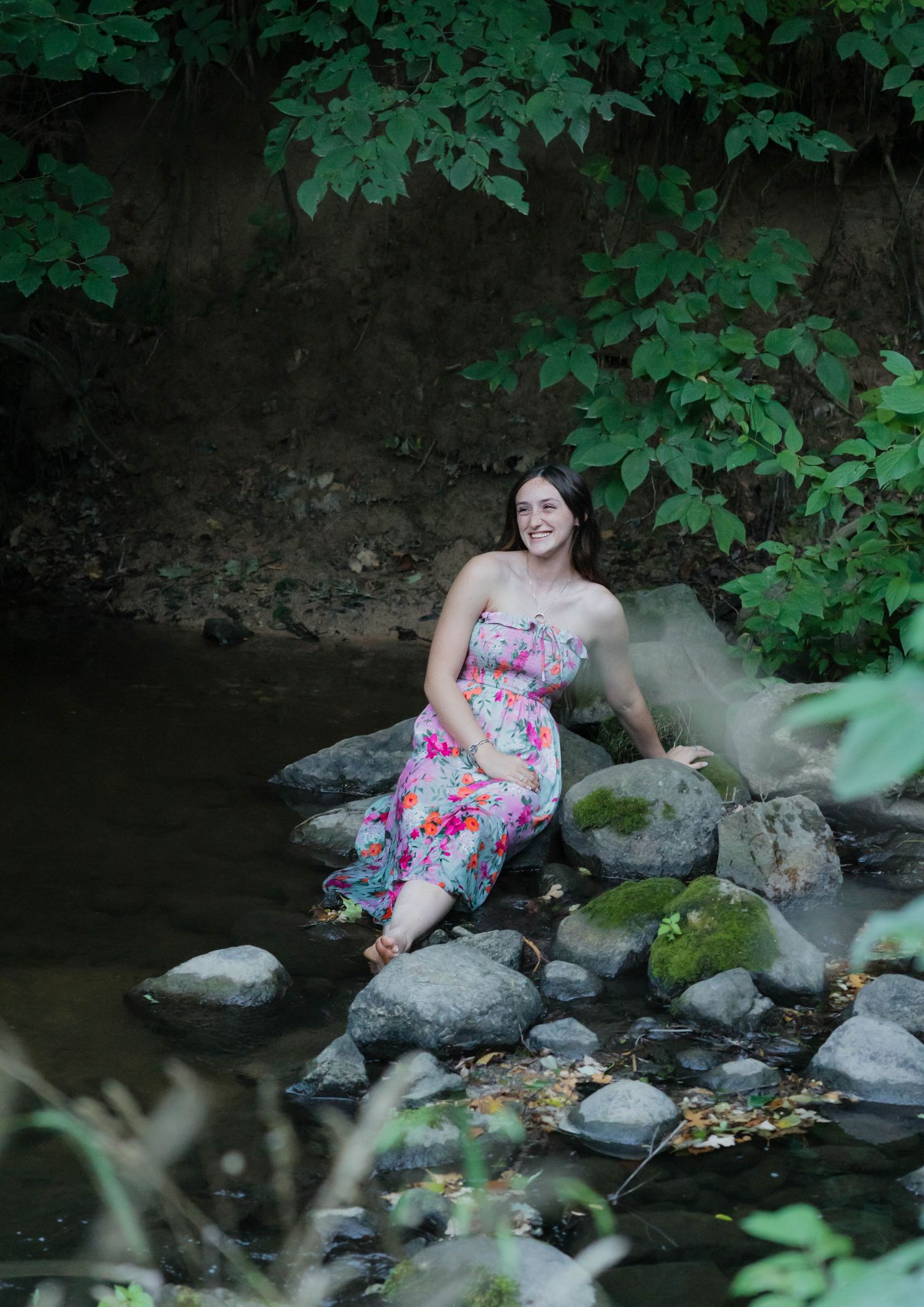 Smiling young woman in floral dress sitting on mossy rocks beside a peaceful forest stream.
