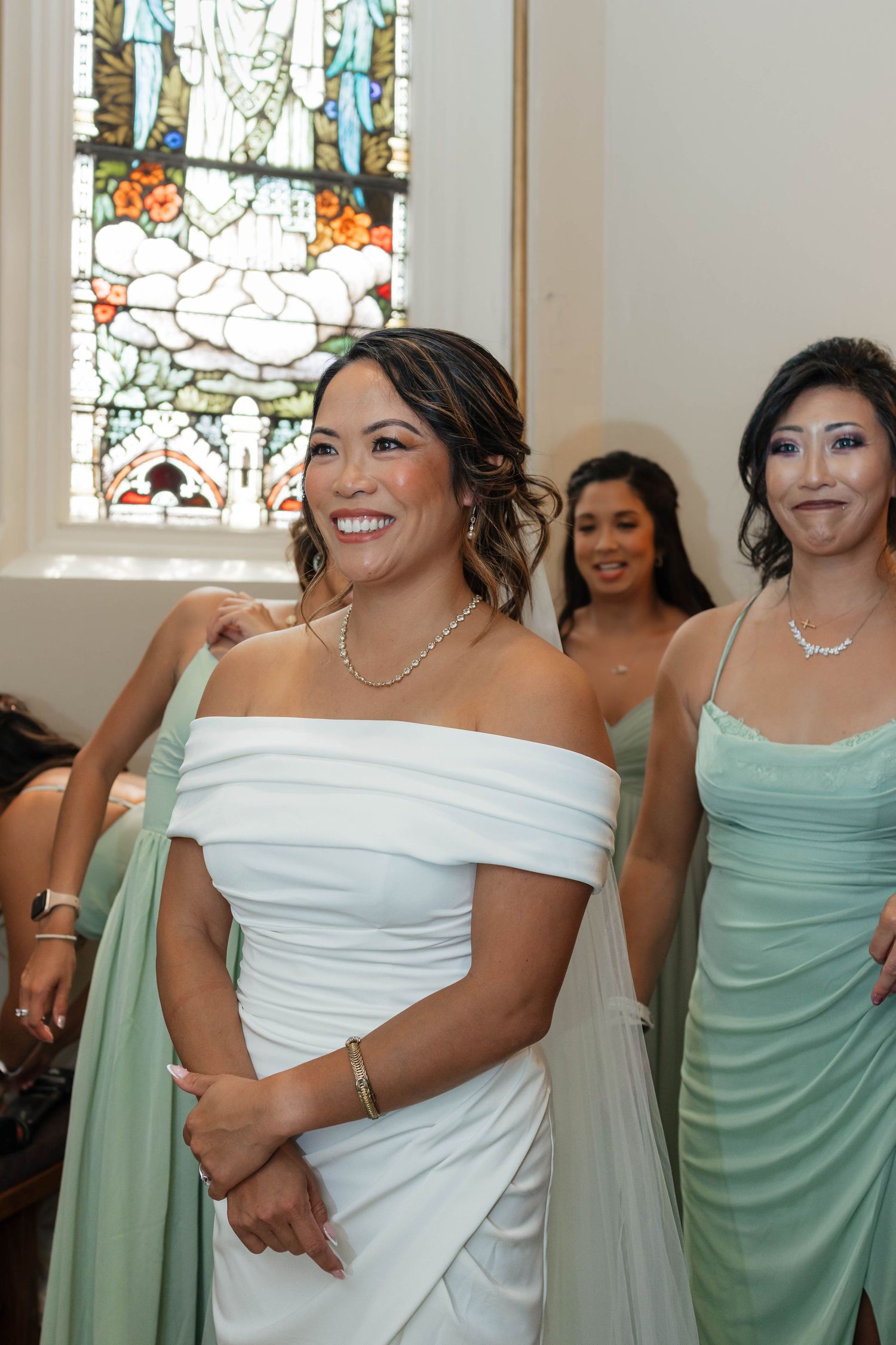 Smiling bride in white off-shoulder gown walks down aisle with bridesmaids in sage green dresses.