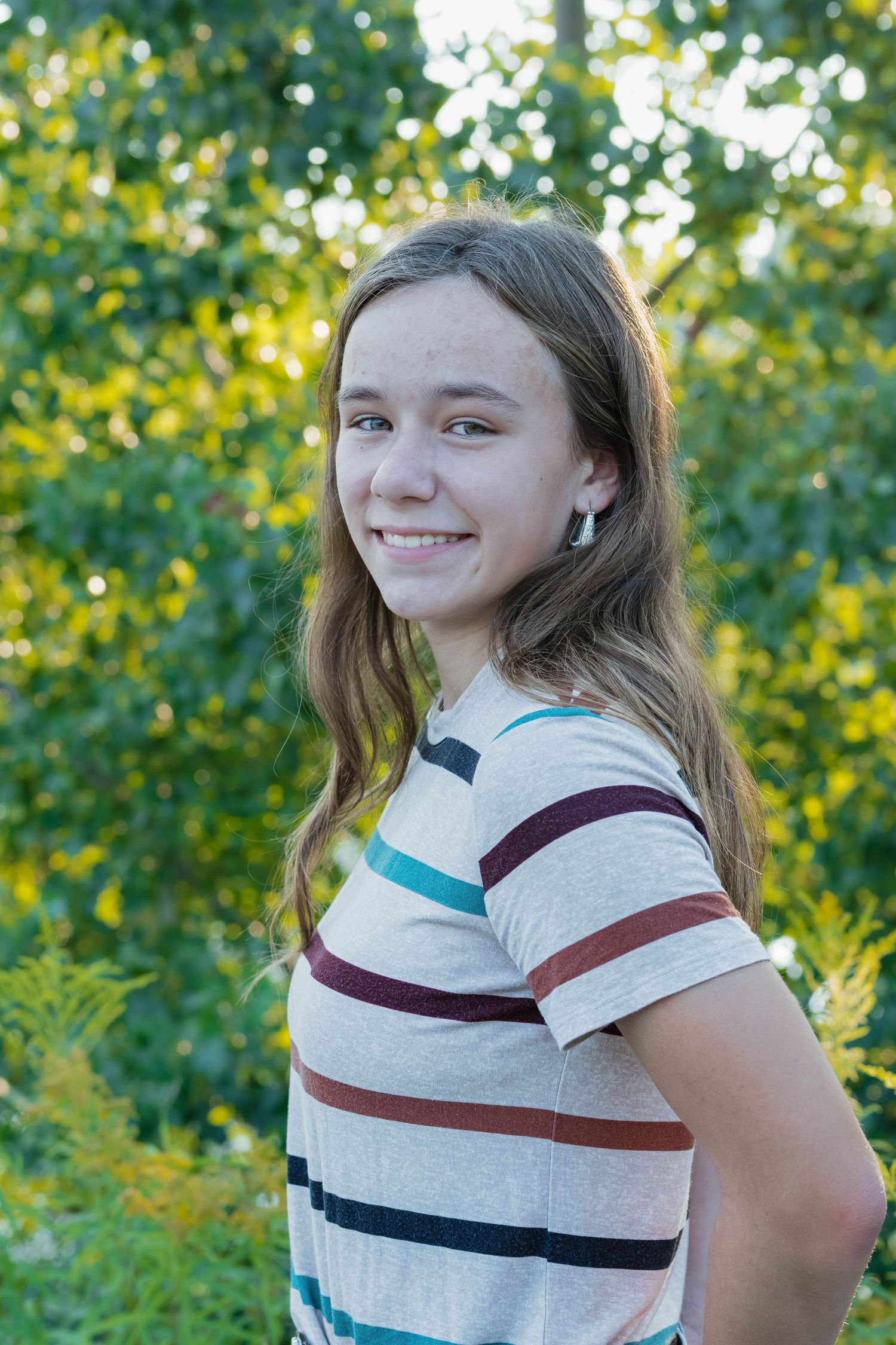 Smiling teenage girl with long brown hair wearing a striped t-shirt outdoors with green trees in background.