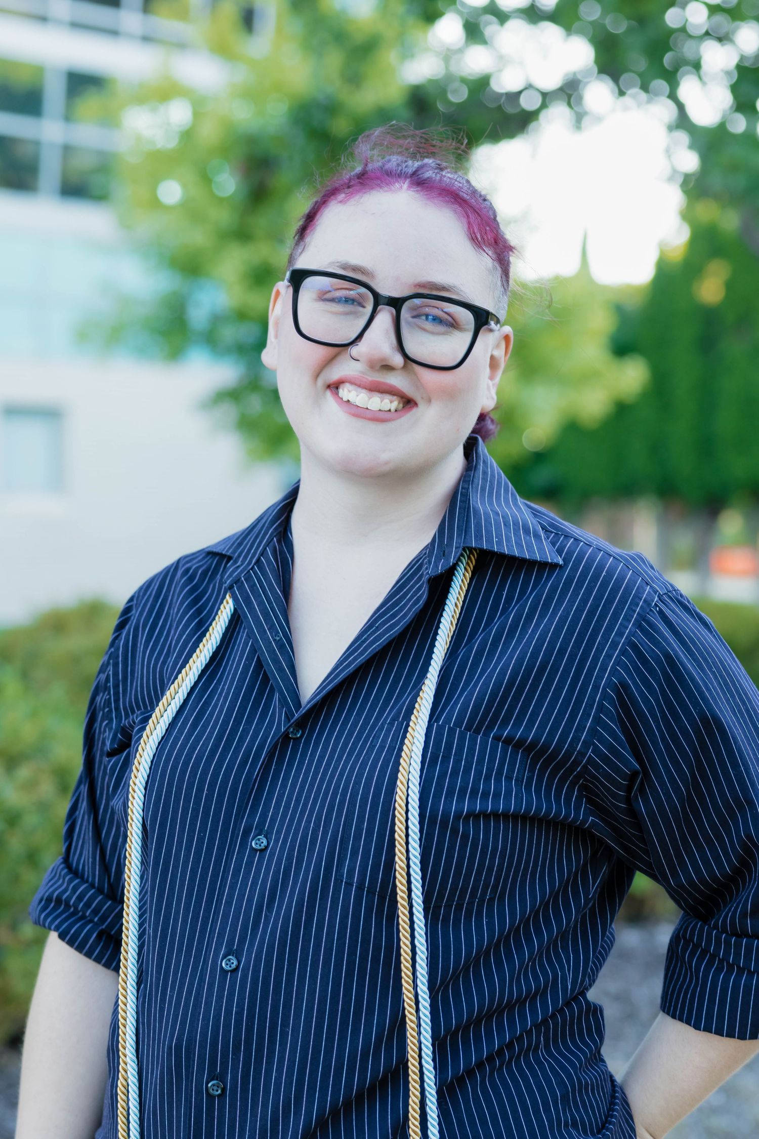 Smiling graduate with pink hair and glasses wearing striped shirt with gold honor cords.