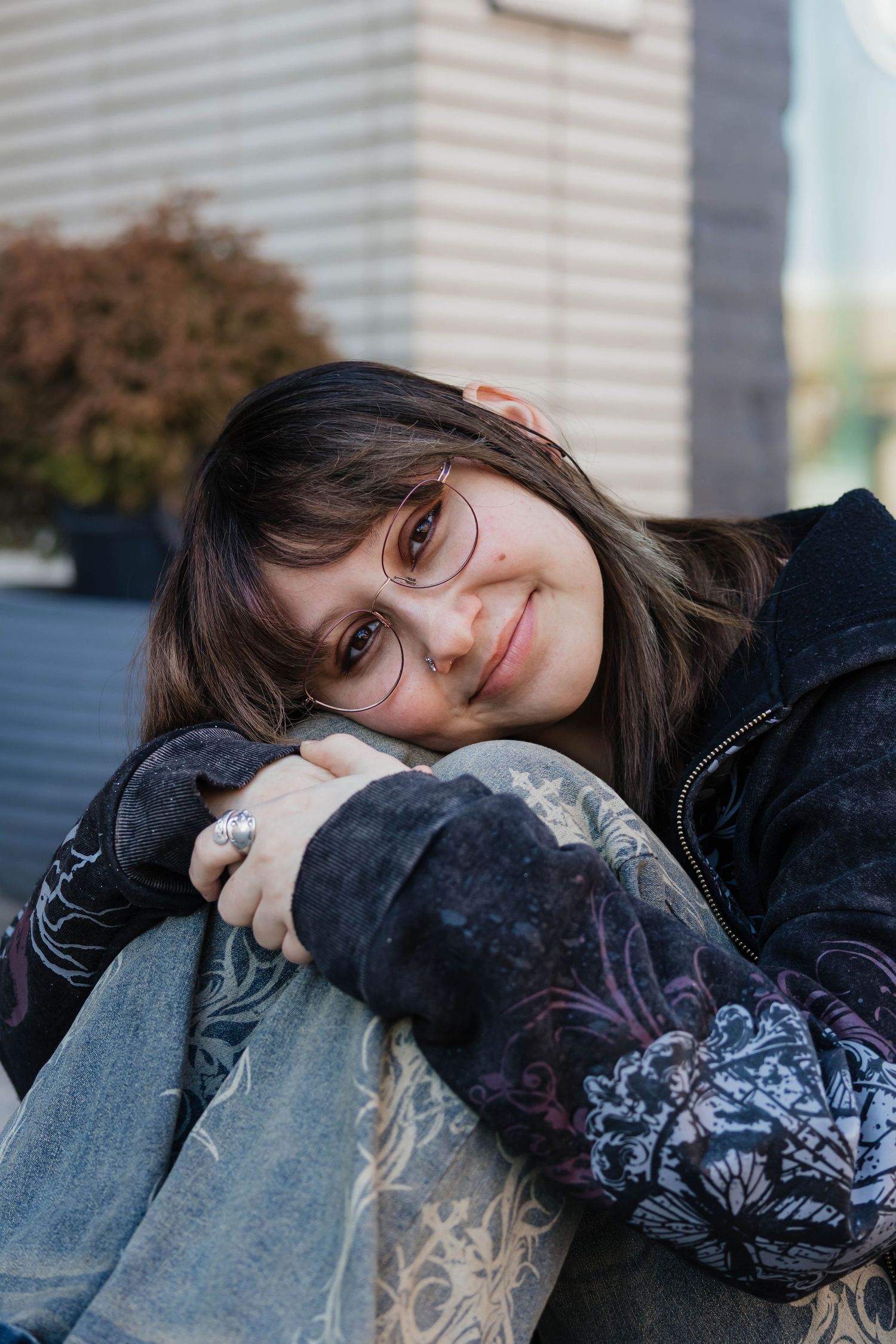 Young woman with glasses resting her chin on her knees, smiling softly outdoors in casual boho style.