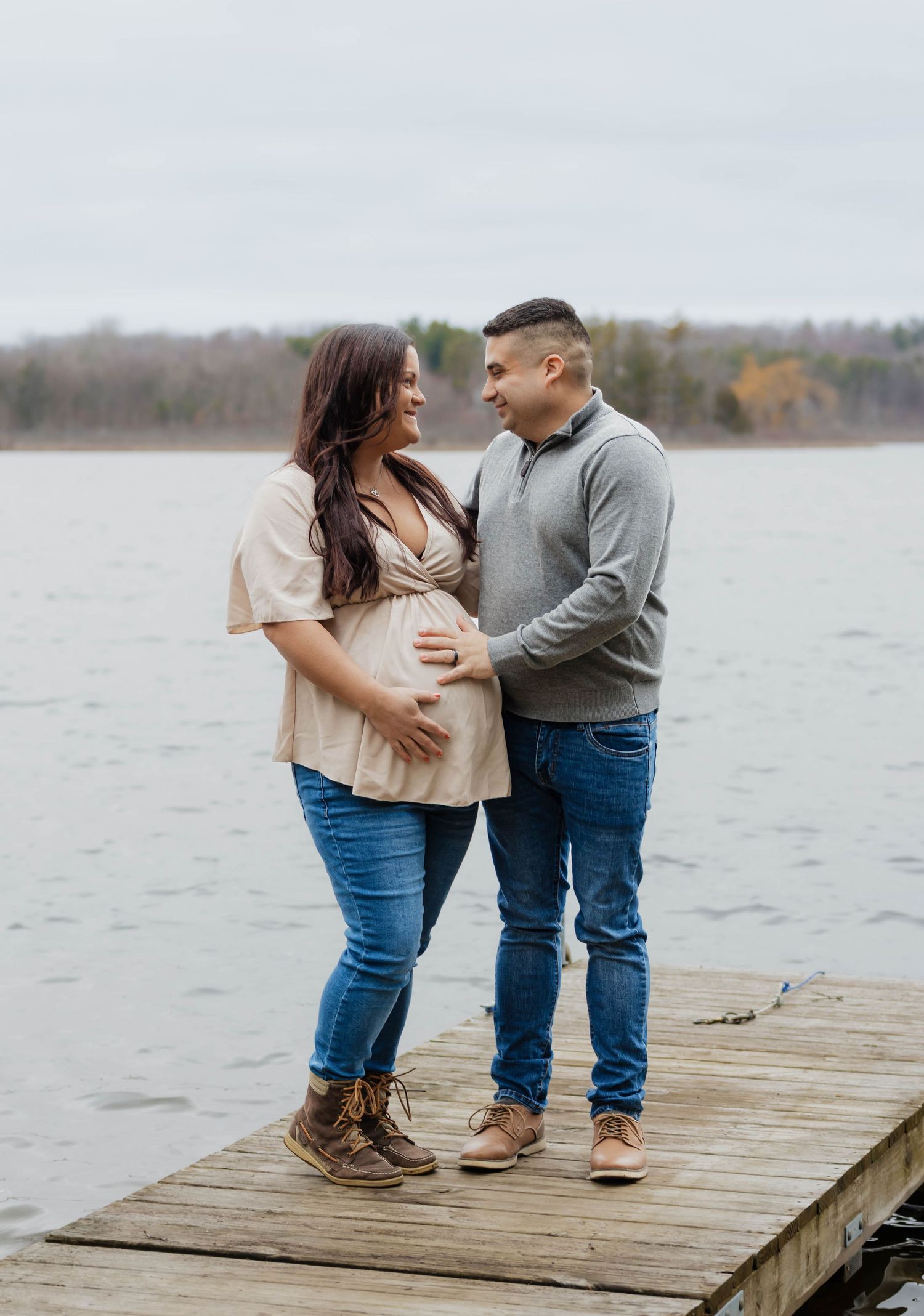 Happy expecting couple standing on a wooden dock by a lake, cradling baby bump during maternity session.