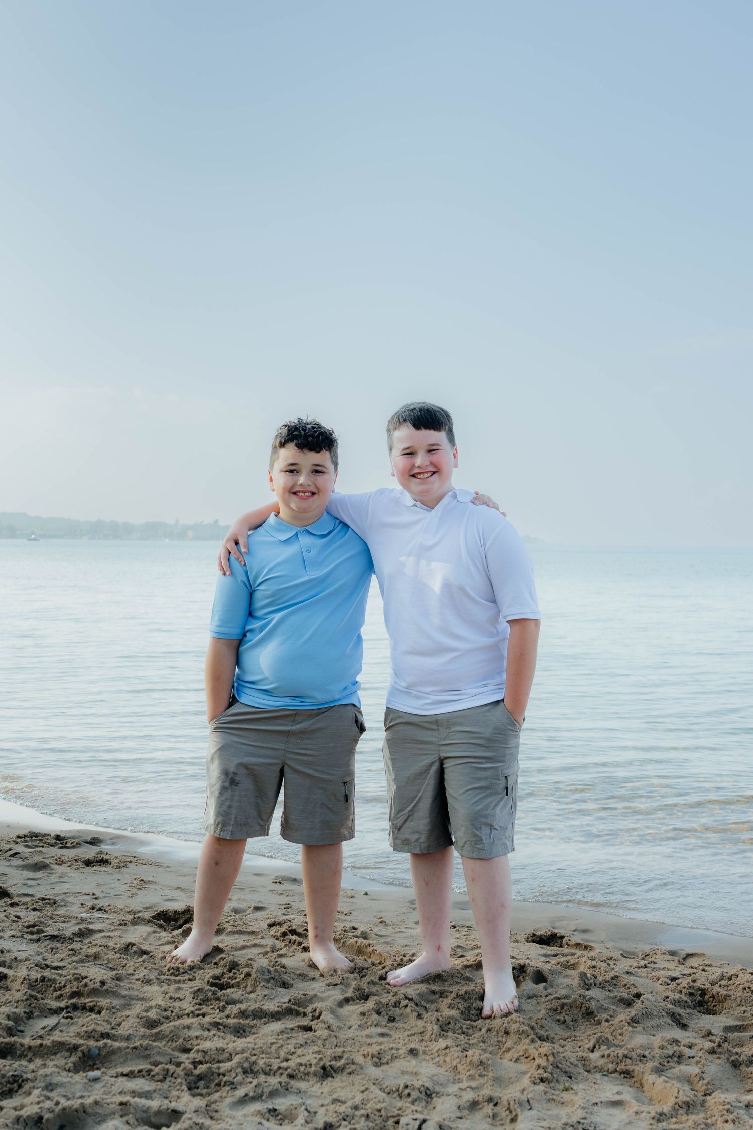 Two boys smiling together on a sandy beach with calm ocean water behind them.