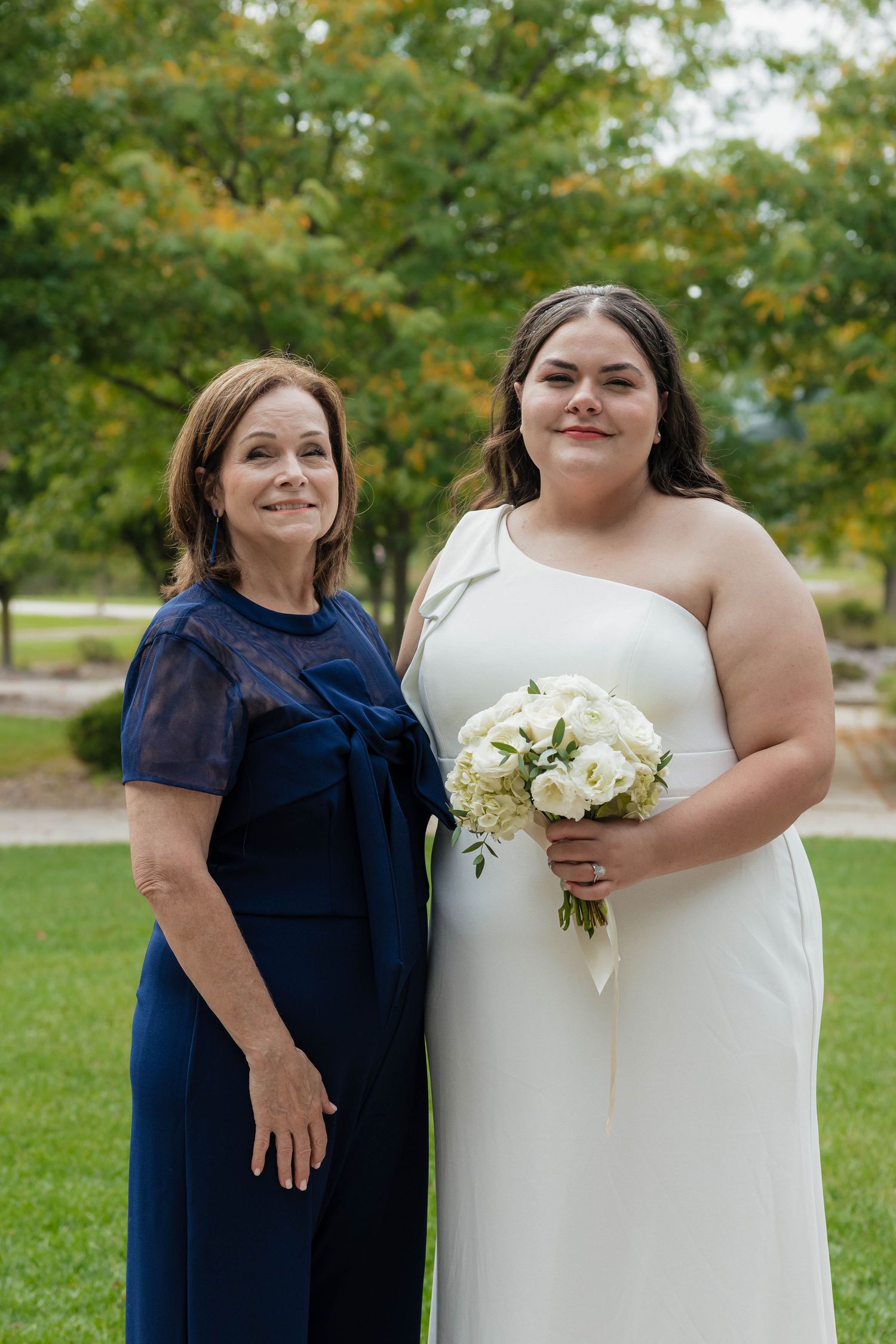 Bride in white one-shoulder gown holds bouquet while posing with older woman in navy dress.