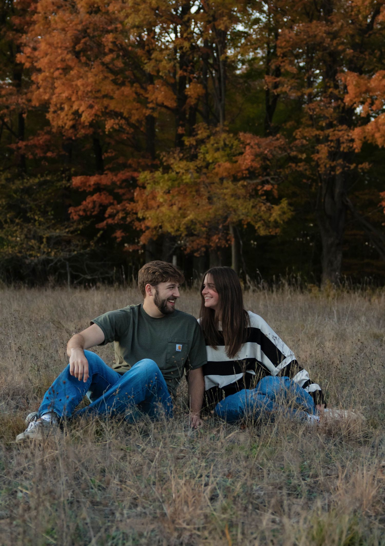 Couple sitting in a field of dry grass with colorful autumn trees behind them.