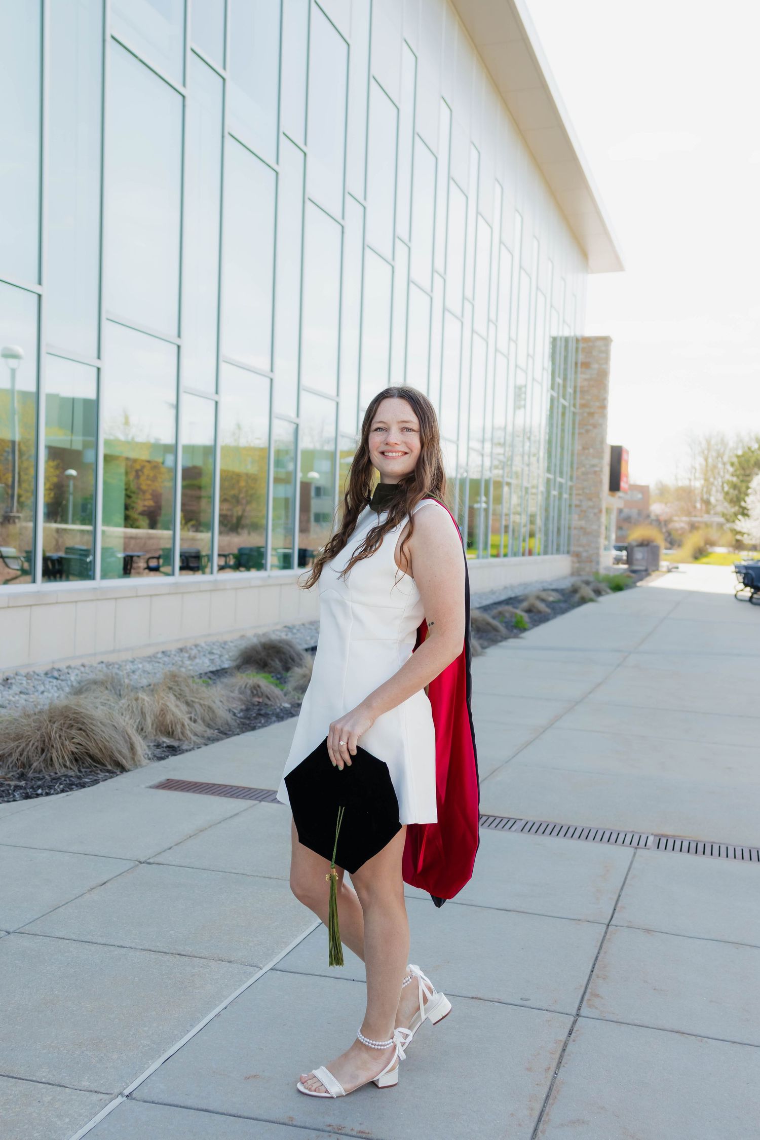 Stylish graduate in white dress holding black cap with red hood outside modern university building.