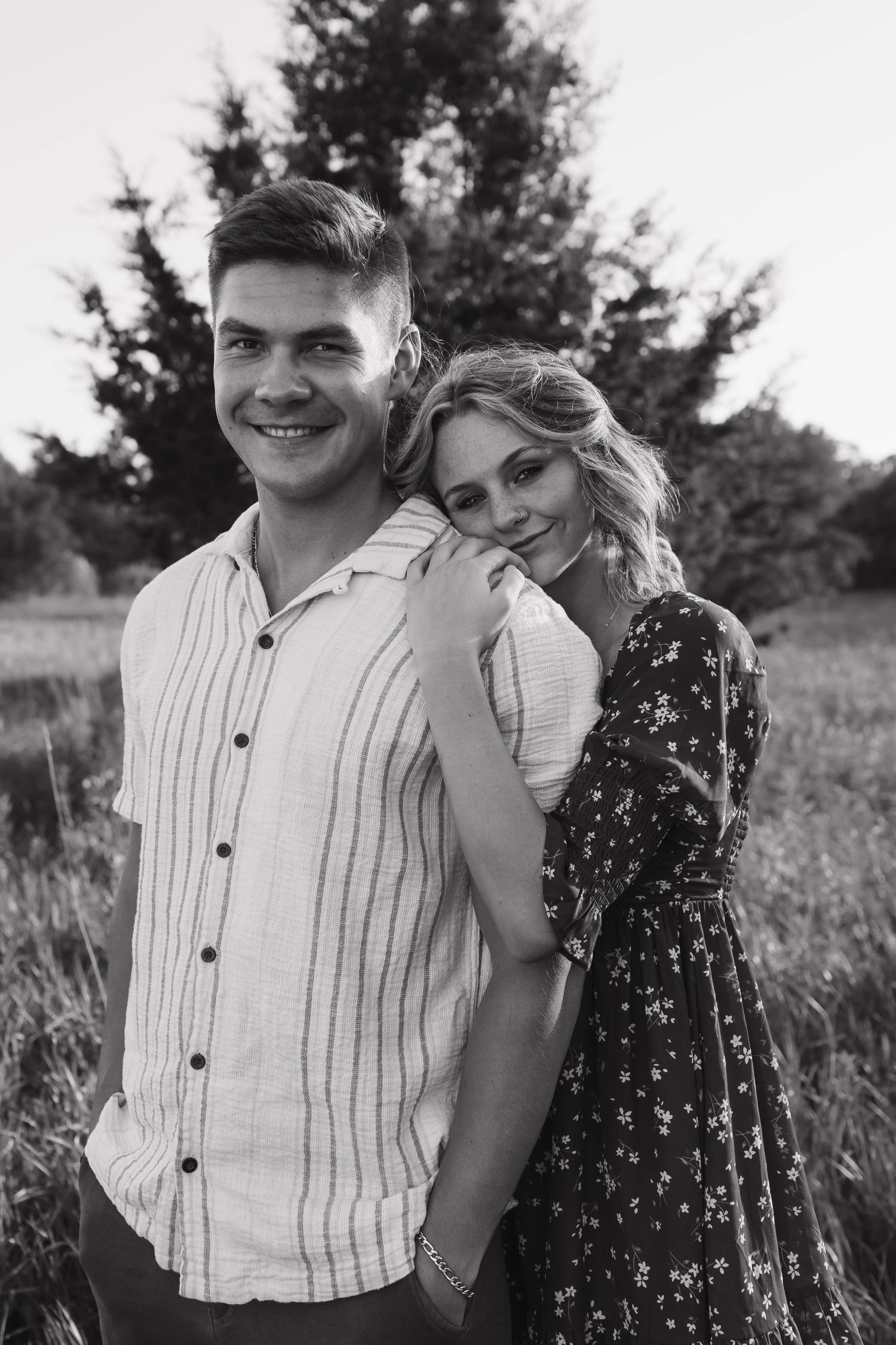 Black and white photo of smiling couple outdoors, woman in floral dress leaning on mans shoulder affectionately.