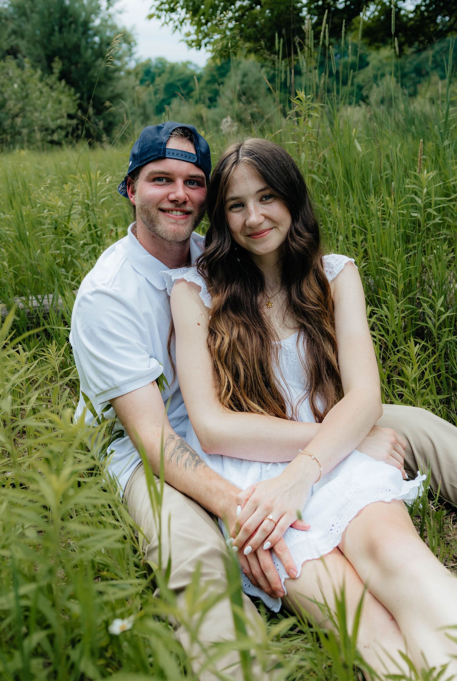 Smiling engaged couple sitting in tall green meadow grass, woman in white dress showing engagement ring.