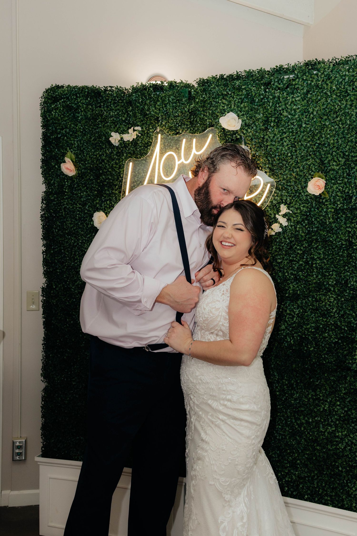 Happy bride and groom posing in front of a green floral wall with a glowing neon sign.