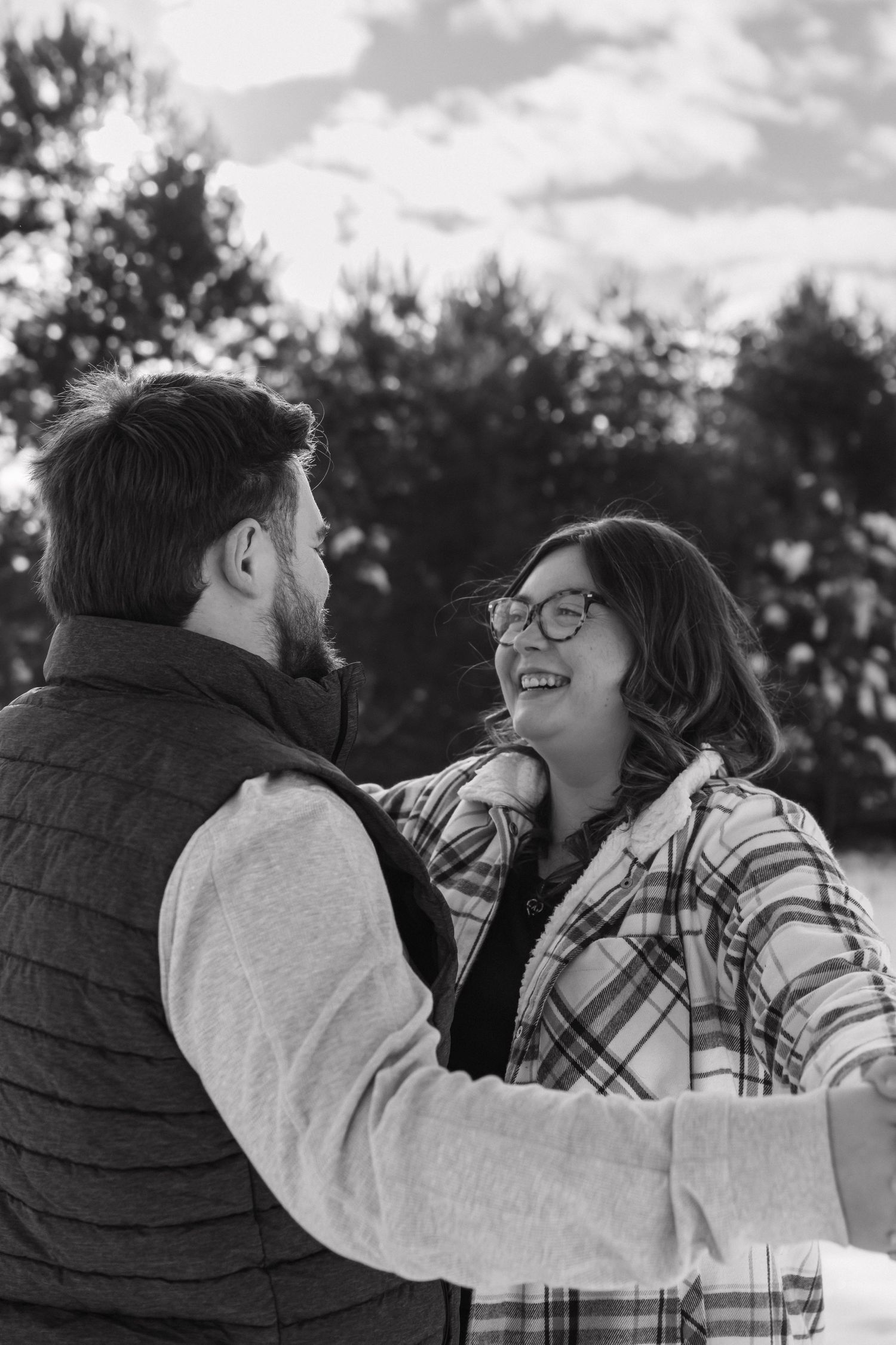 Black and white photo of a laughing couple embracing outdoors in winter, woman wearing plaid jacket and glasses.