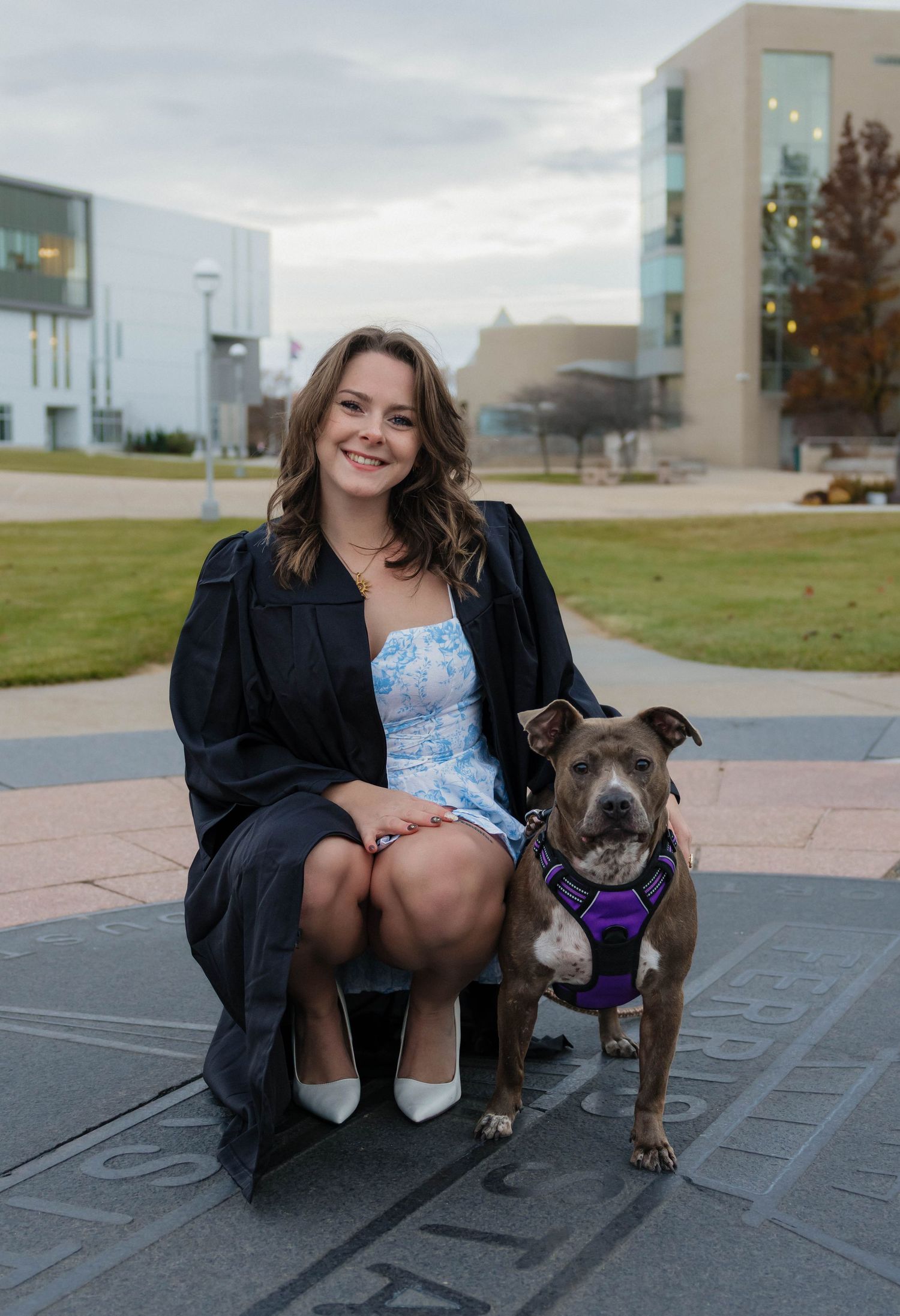 Smiling graduate in cap and gown posing with a gray pit bull on a university campus.