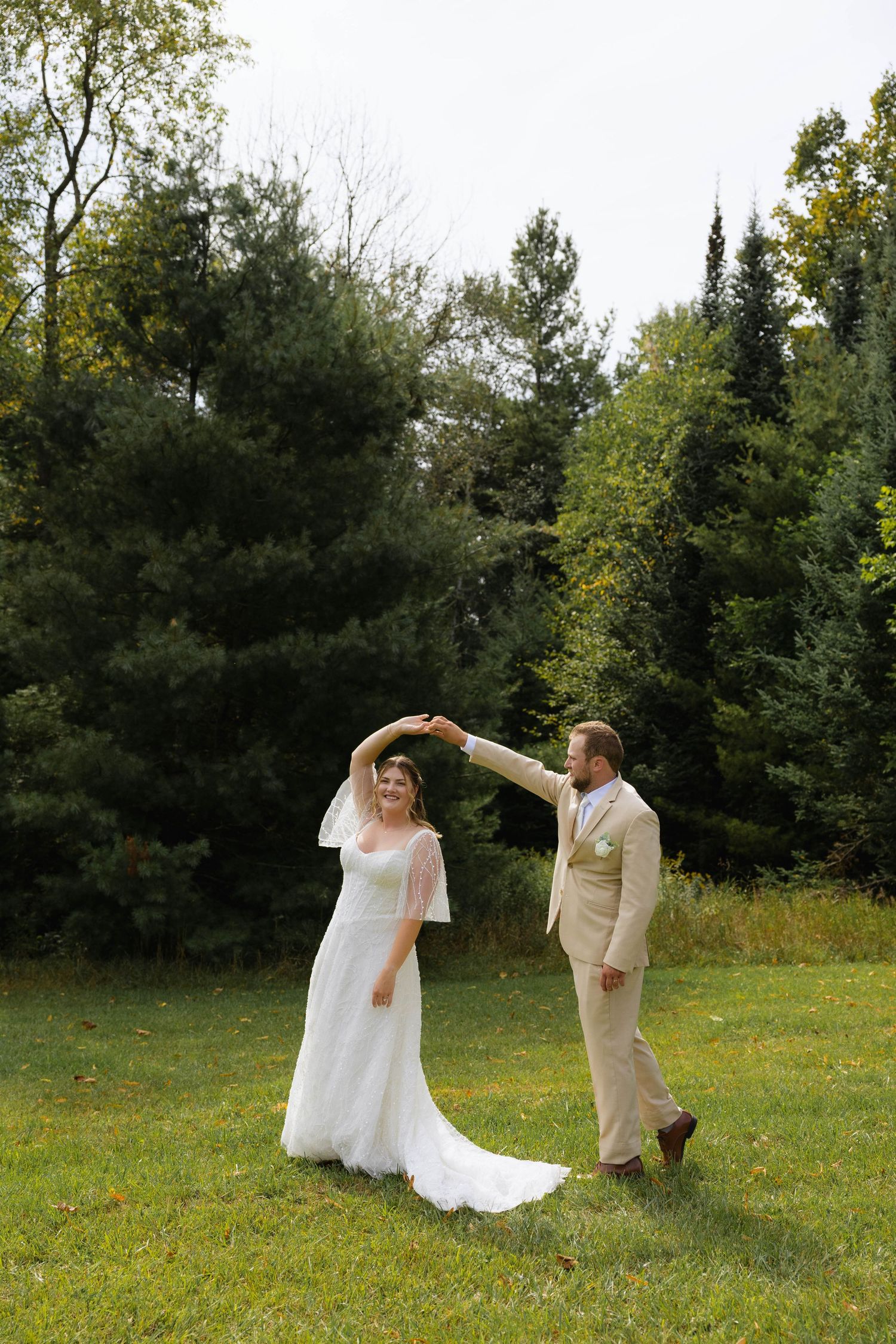 Bride and groom dancing outdoors on green grass with lush evergreen trees in background during wedding photo session.