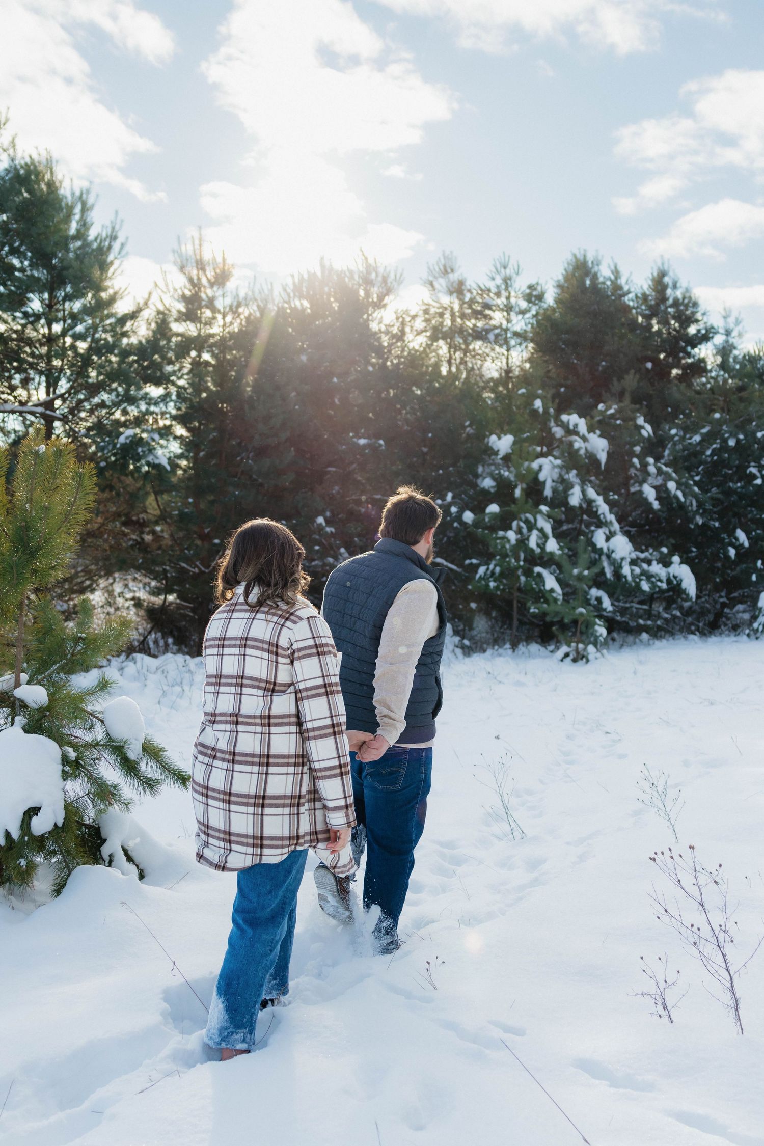 Couple holding hands walking through a snowy pine forest path with sunlight streaming through trees.