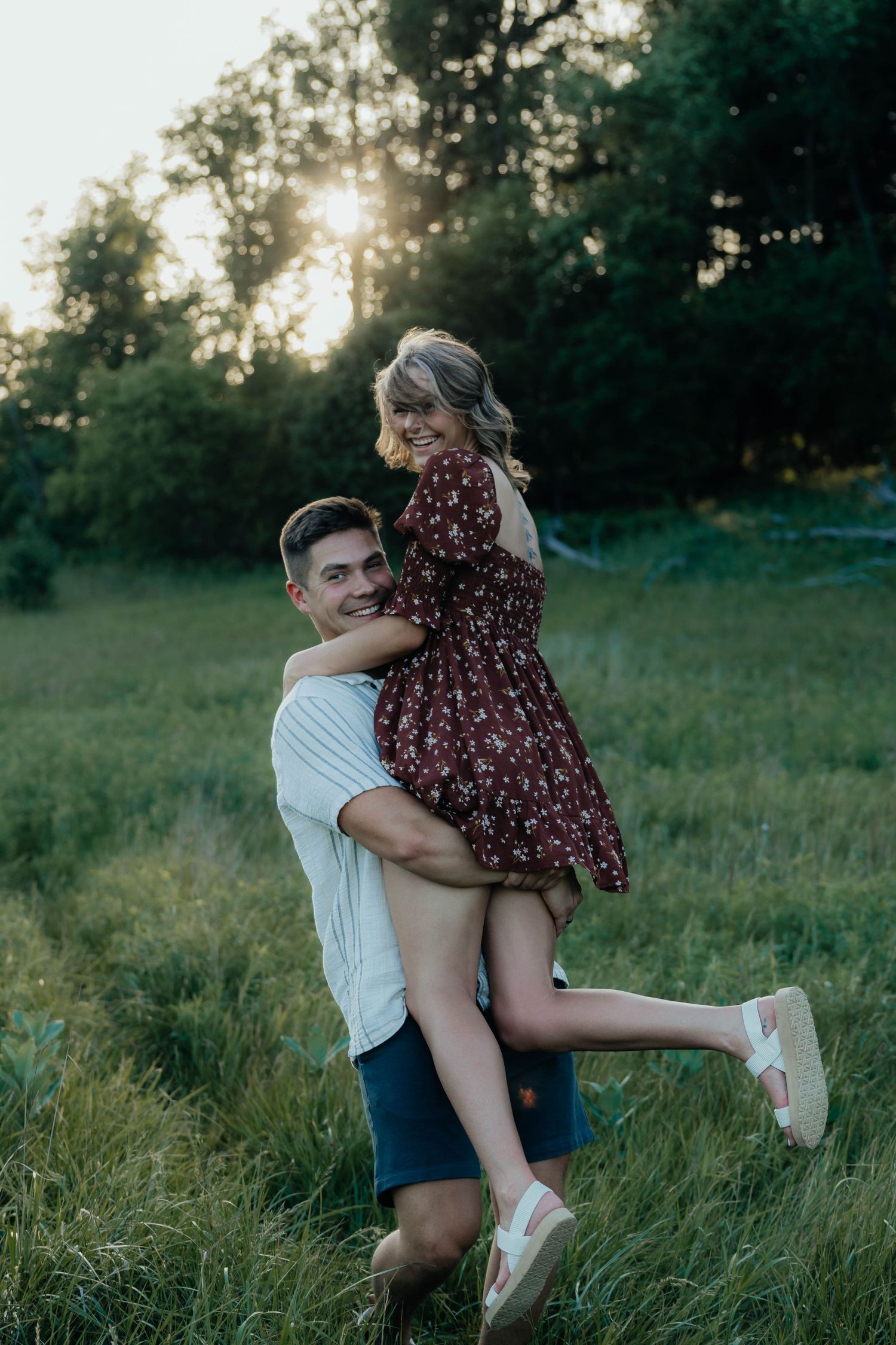 Couple sharing a joyful piggyback moment in a sunlit green meadow, woman in floral dress laughing.