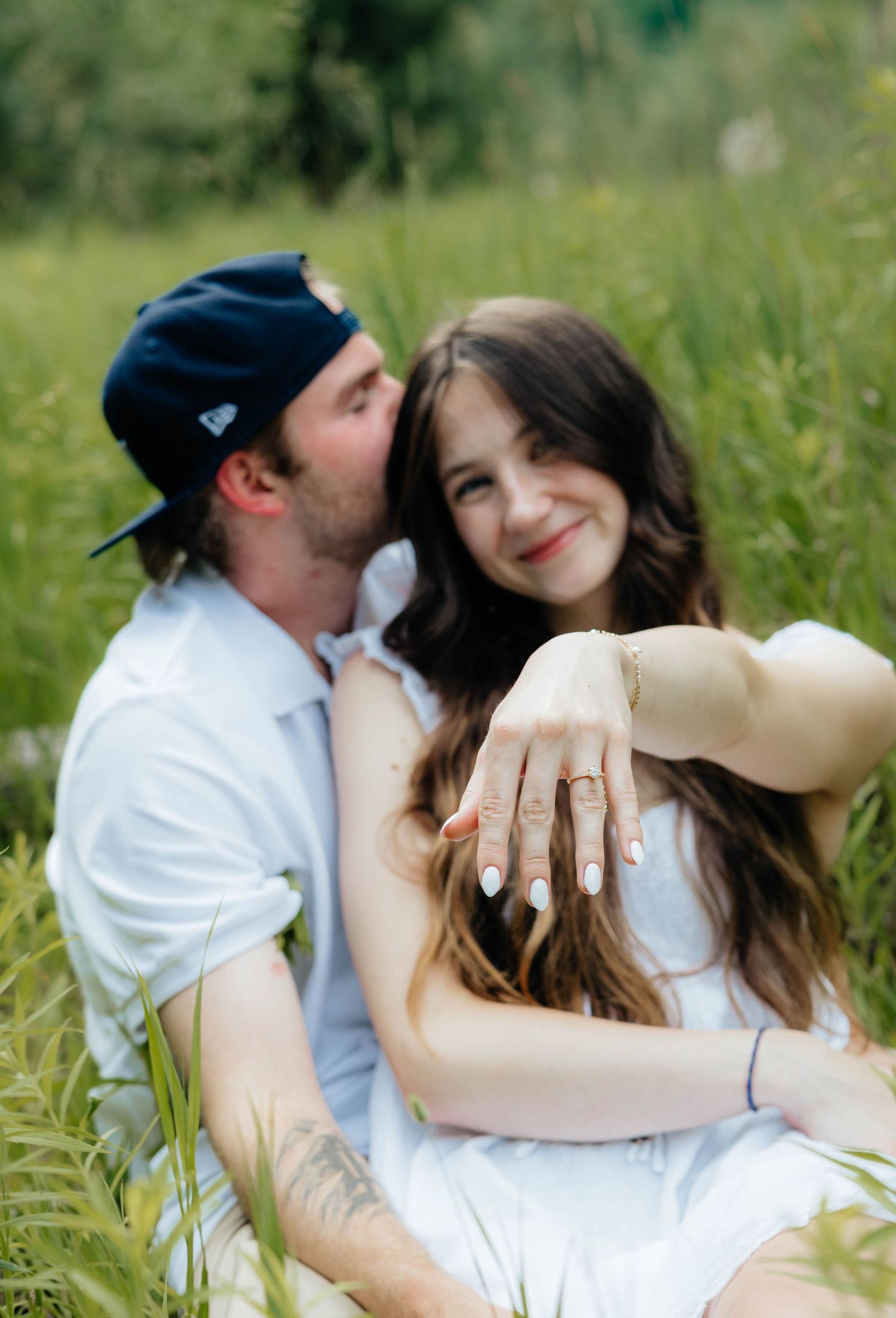 Happy woman showing off engagement ring while partner kisses her cheek in a lush green field.