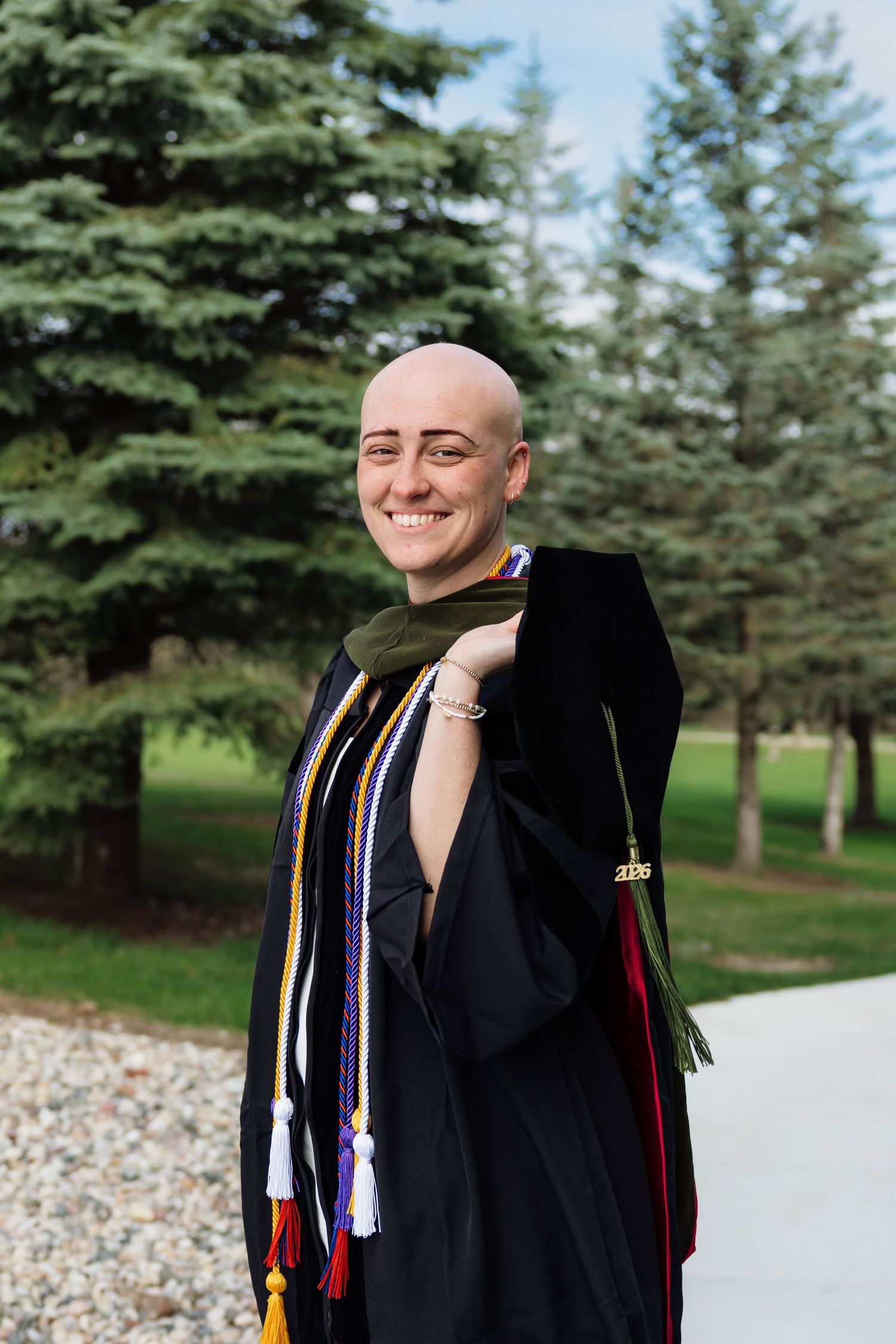 Confident bald woman smiling in black doctoral graduation gown with colorful honor cords outdoors.