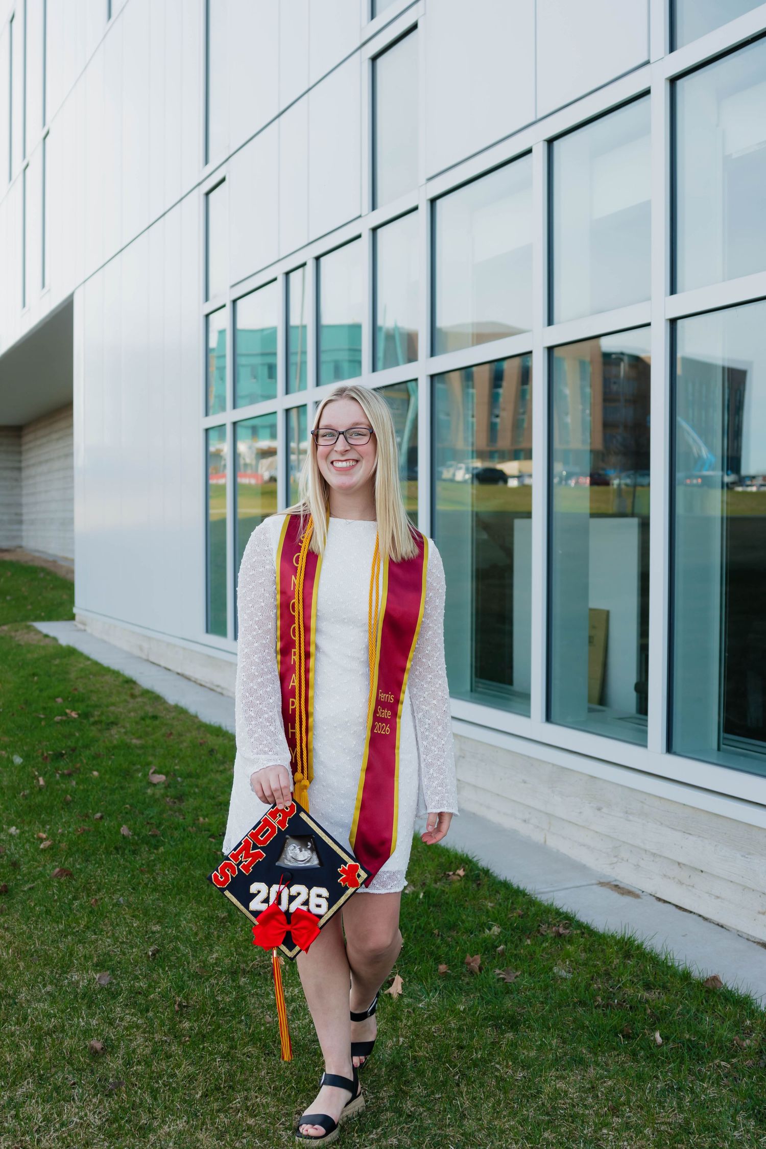 Female Ferris State 2026 graduate in white dress with maroon stole holds decorated graduation cap outdoors.
