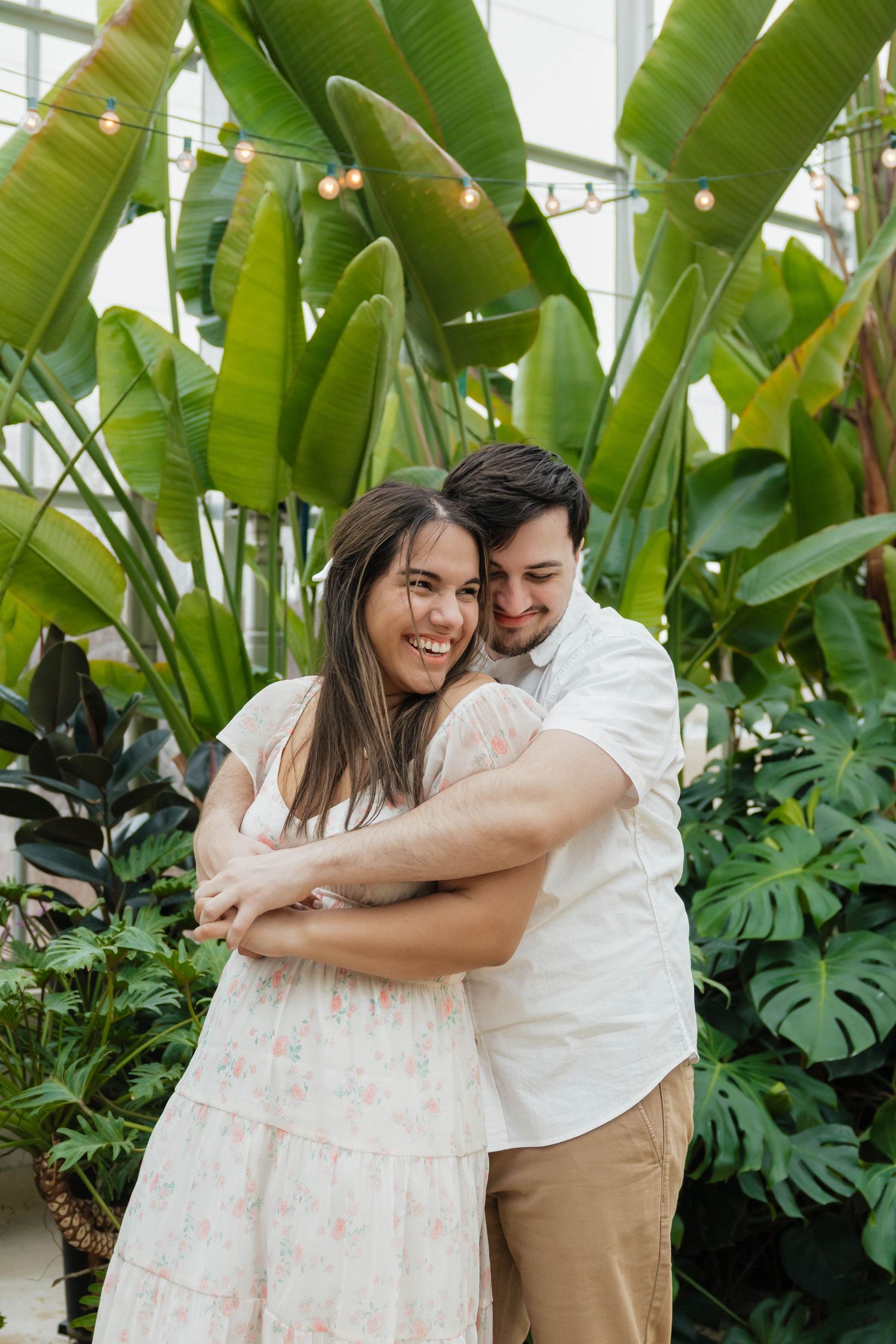 Couple embracing and smiling among tropical plants in a greenhouse engagement photo.